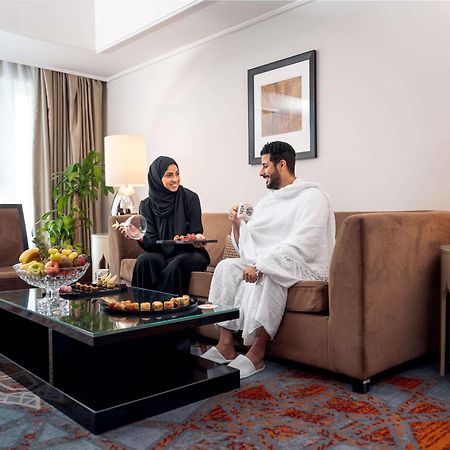 Swissotel Makkah Mecca Exterior photo The photo depicts a comfortable living space where a man and woman are seated together. The man is wearing traditional attire, while the woman is dressed in a black abaya. They appear to be engaged in a pleasant conversation. A coffee table in front 