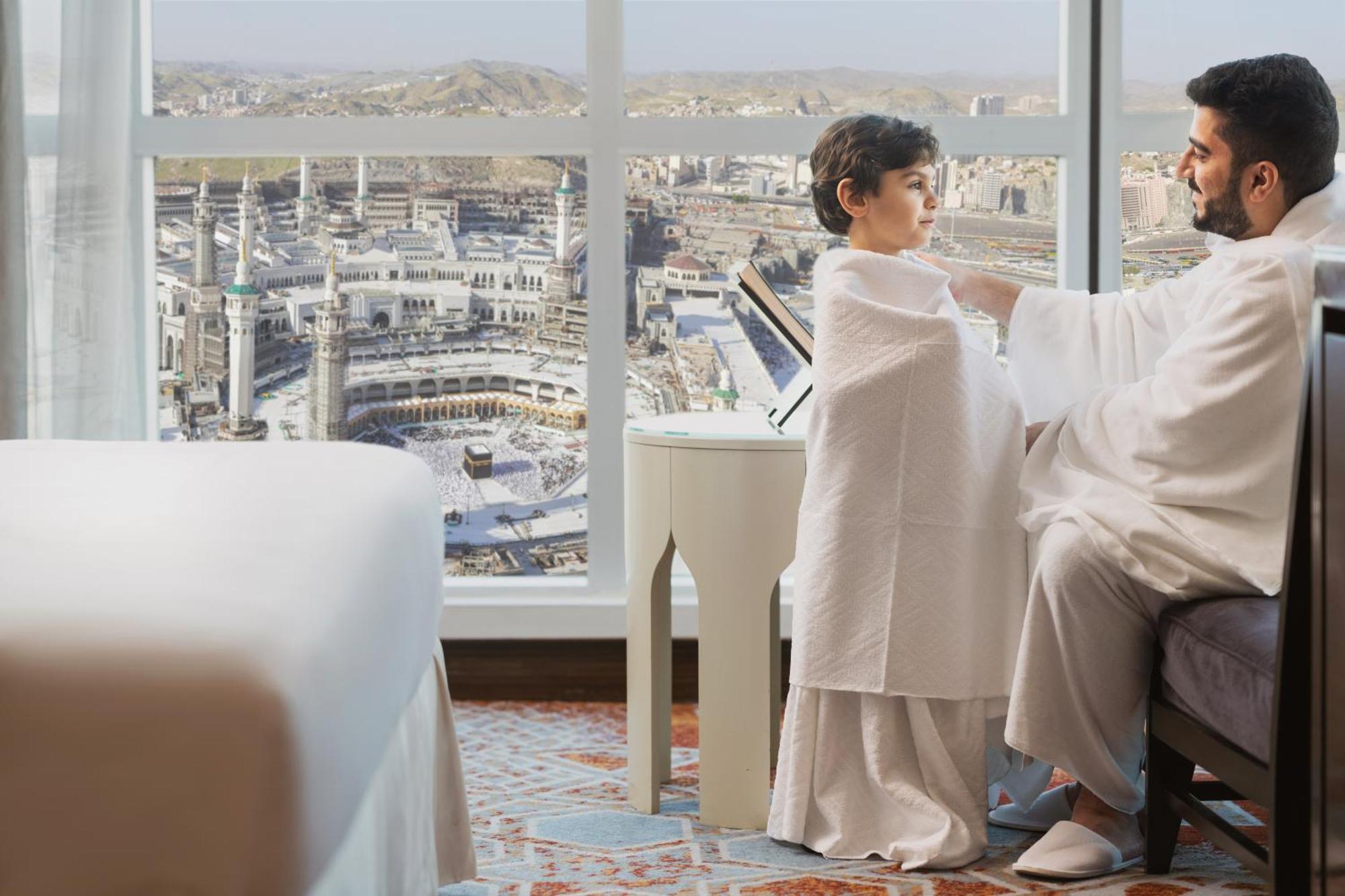 Swissotel Makkah Mecca Exterior photo The photo depicts a father and son sitting together in a hotel room. The father is wrapping a cloth around the boy, who appears to be preparing for a religious pilgrimage or ceremony, likely in the context of Hajj or Umrah. The background showcases a