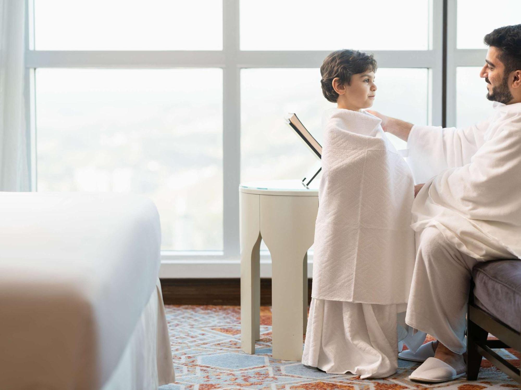 Swissotel Makkah Mecca Exterior photo The photo shows a man and a child in a bright, contemporary hotel room. The man, dressed in traditional attire, is gently assisting or guiding the child, who is also wrapped in a similar garment. They are seated near a small table by a large window, 