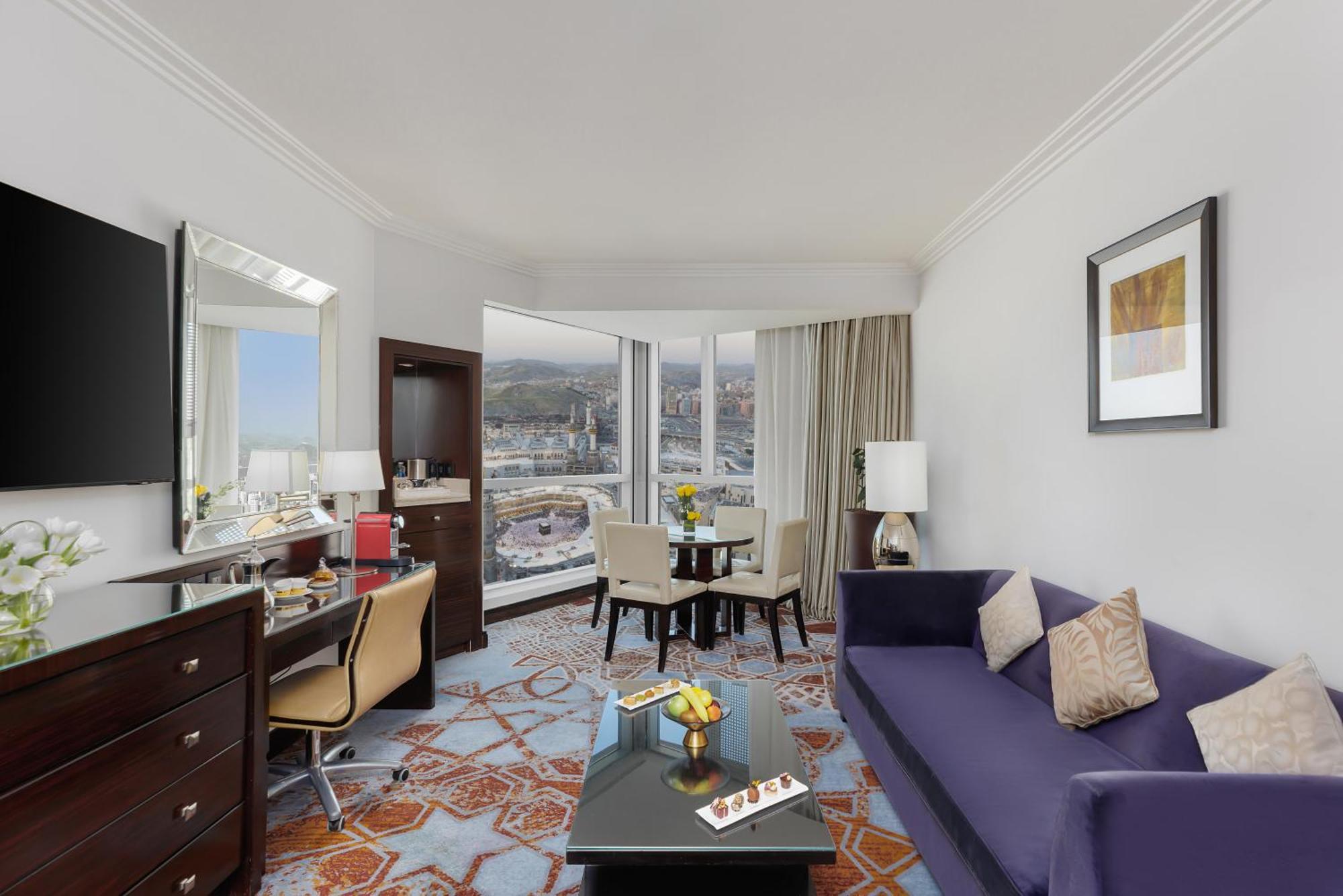 Swissotel Makkah Mecca Exterior photo The photo shows a hotel room with a modern and stylish design. There is a comfortable purple sofa in the foreground, facing a small coffee table with some decorative items on it. Near the sofa, there is a work desk with a chair and a flat-screen TV m