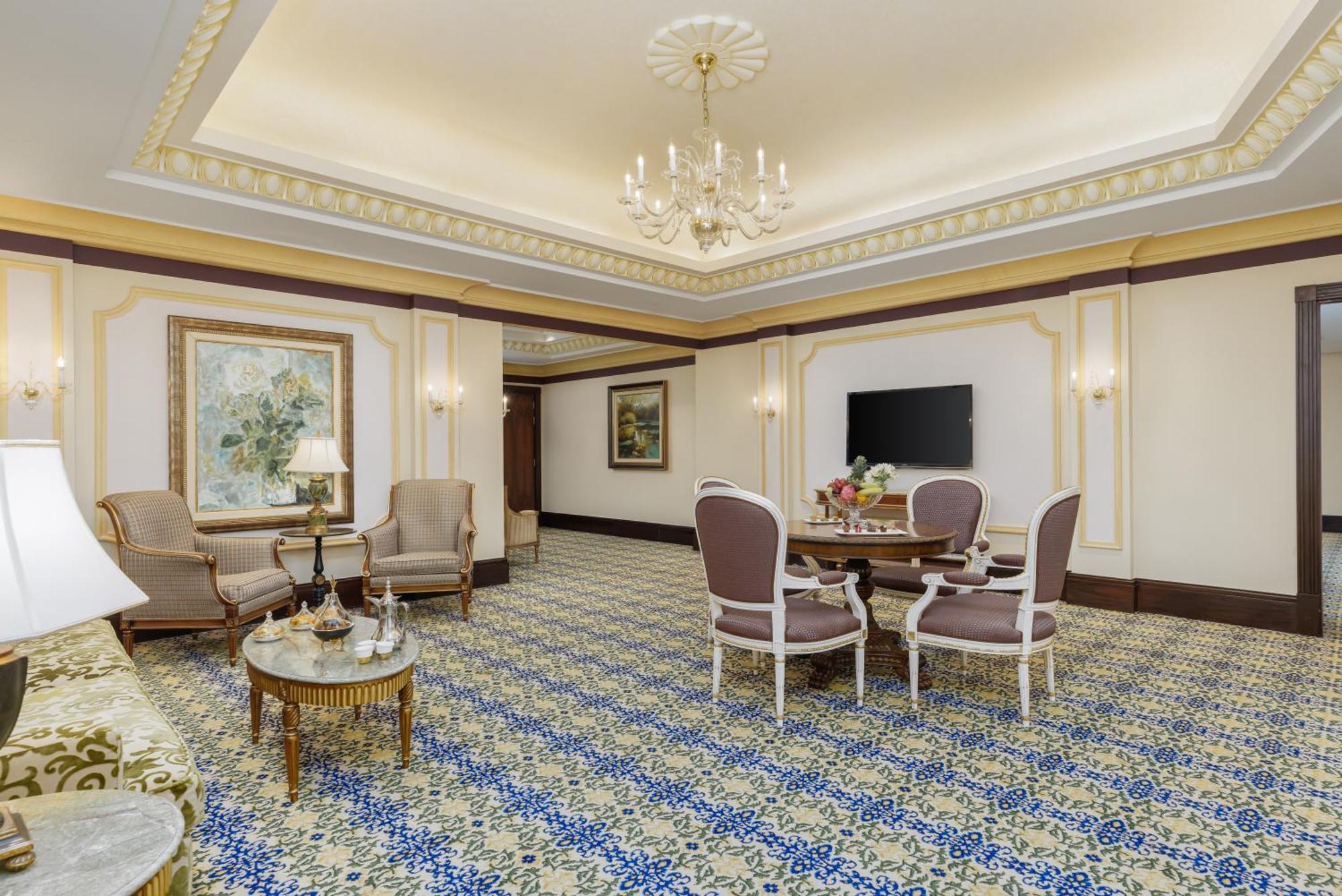 Swissotel Makkah Mecca Exterior photo The photo shows a luxurious interior space, likely a hotel suite or a high-end lounge area. It features a beautifully patterned carpet with blue and cream tones. The room is furnished with two elegant armchairs and a round dining table surrounded by 