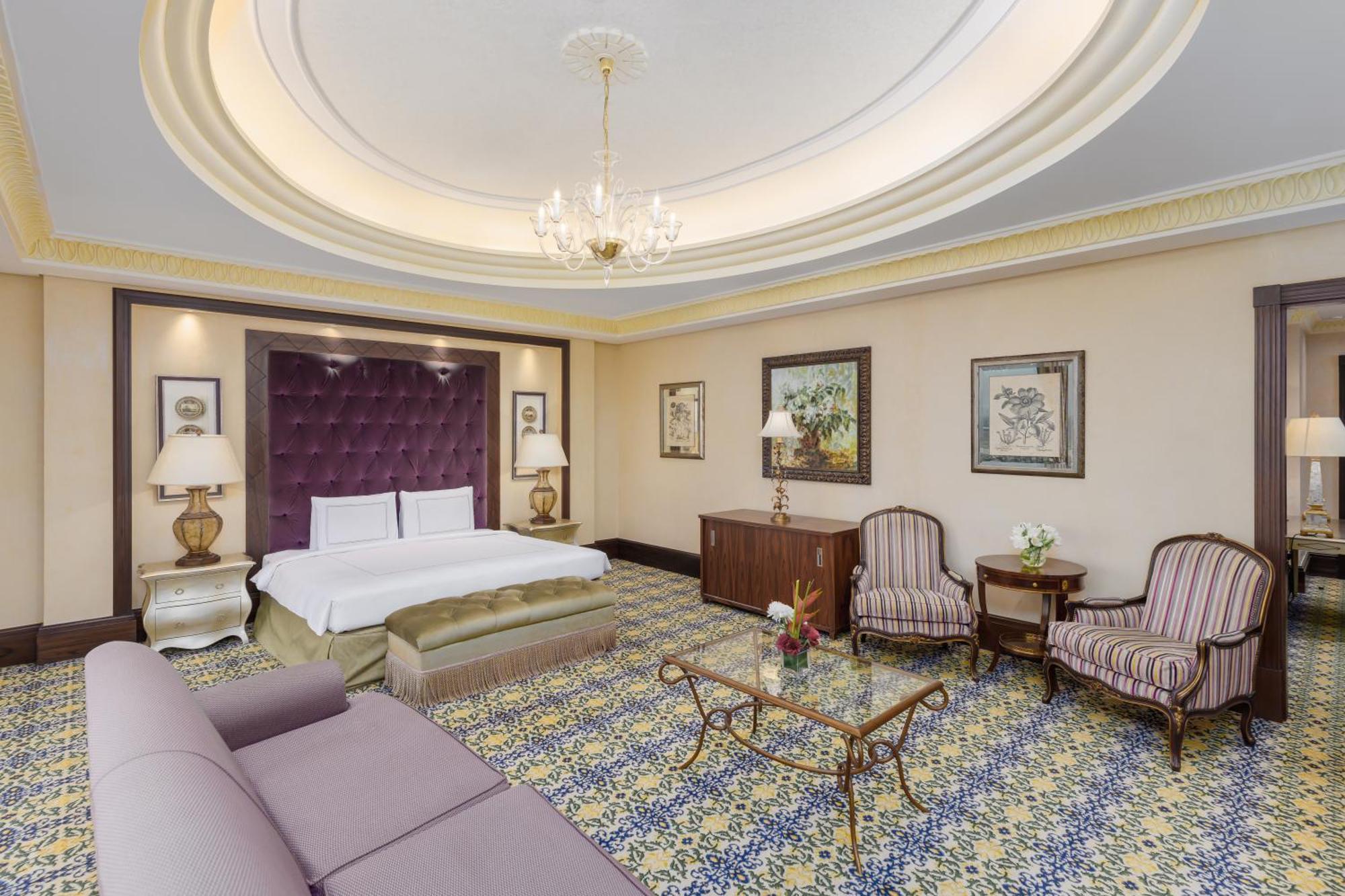 Swissotel Makkah Mecca Exterior photo The photo shows an elegantly designed hotel room. It features a large bed with a tufted purple headboard, complemented by two bedside lamps. There is a stylish sofa along one side, offering a comfortable seating area. Across from the bed, there is a 