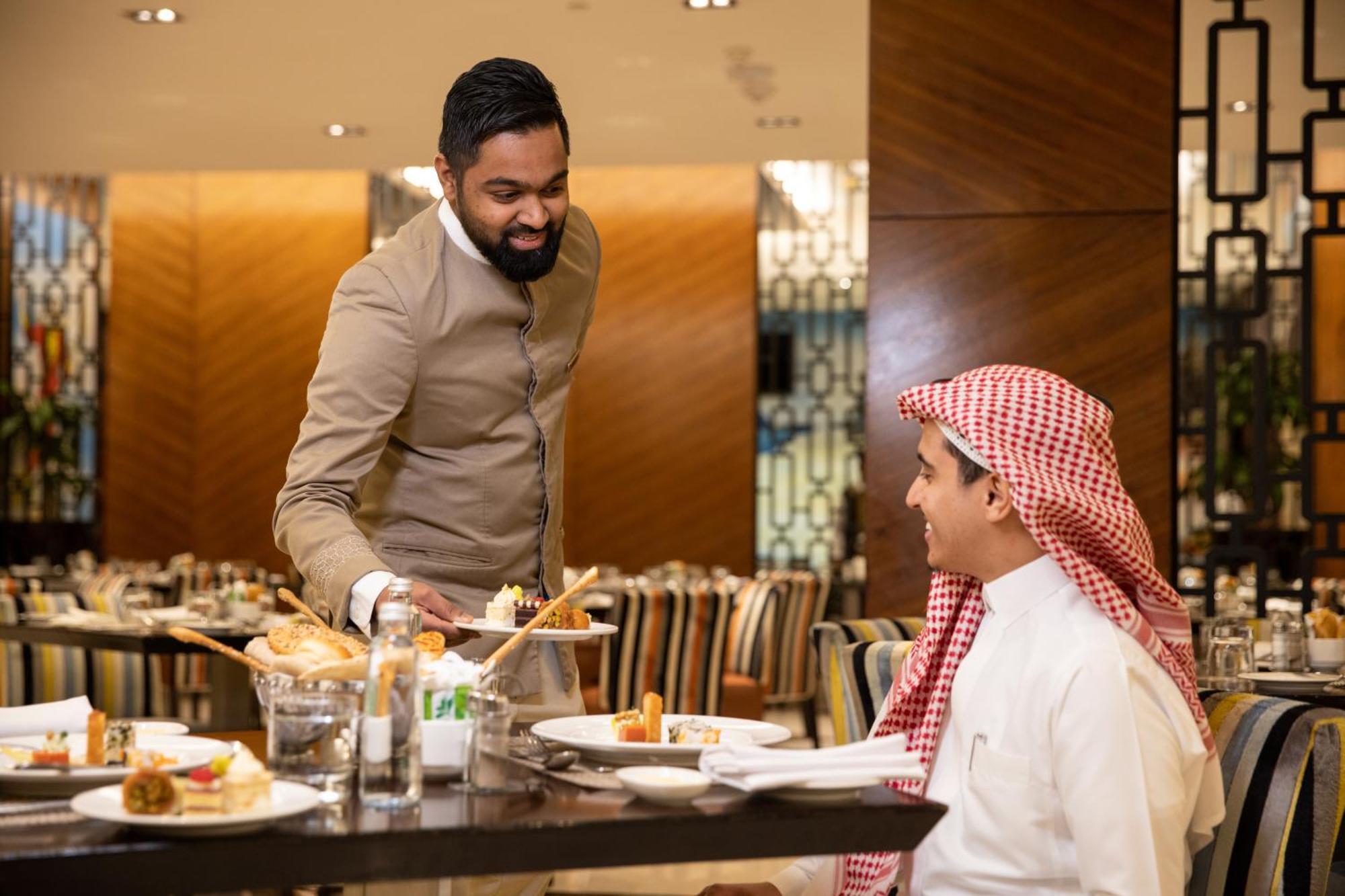 Swissotel Makkah Mecca Exterior photo The photo shows a scene in a restaurant where a waiter is serving a meal to a diner. The waiter, dressed in a formal outfit, appears to be presenting a dish with a smile. The diner, wearing traditional attire, looks engaged and attentive. The dining 