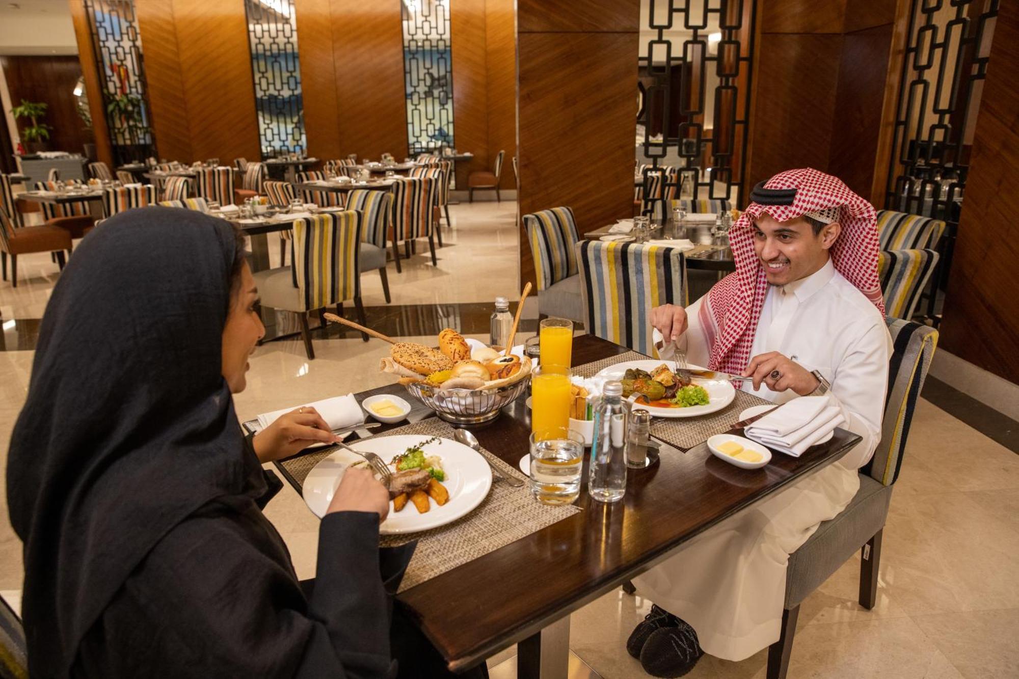 Swissotel Makkah Mecca Exterior photo The photo depicts a dining scene in a restaurant. It features two individuals seated at a table engaging in a meal together. One person is dressed in traditional attire, while the other is wearing a black hijab. The table is set with plates of food, 