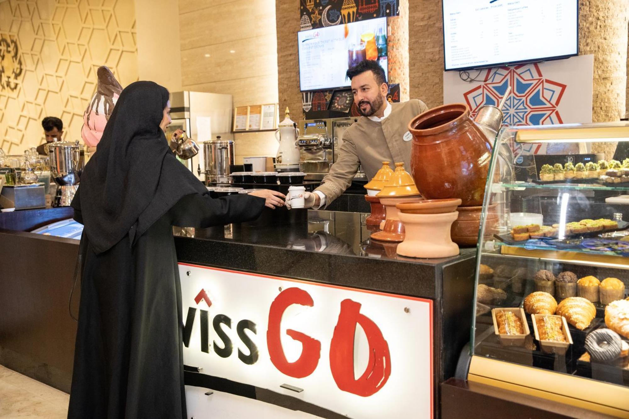 Swissotel Makkah Mecca Exterior photo The photo shows a scene inside a cafe or restaurant. A man in a beige outfit is serving a woman who is wearing a black traditional dress, possibly an abaya. The setting appears modern and includes decorative elements, such as patterned wall decoratio