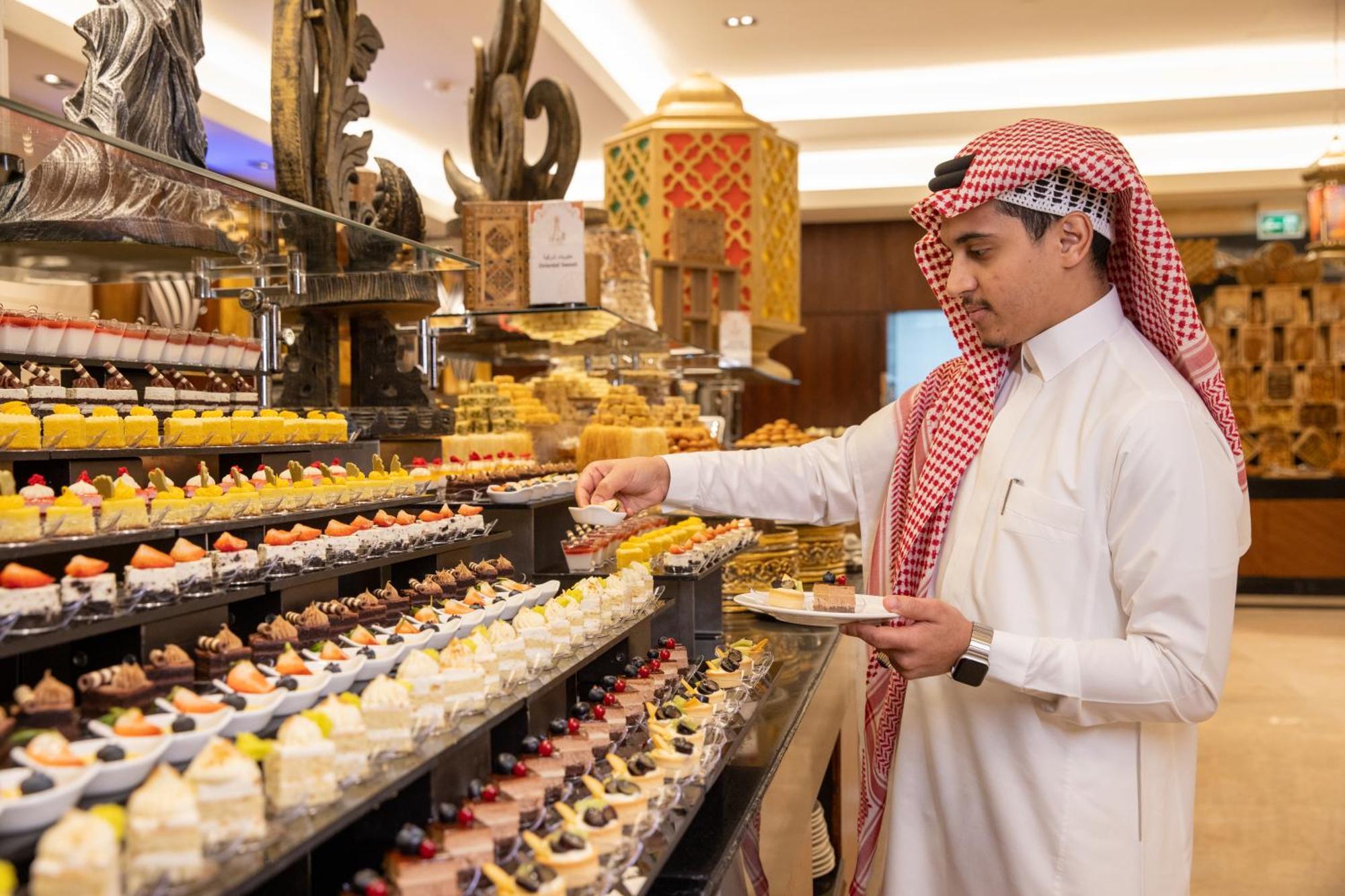 Swissotel Makkah Mecca Exterior photo The photo shows a man dressed in traditional attire, wearing a white thobe and a red-and-white checked ghutrah (headscarf). He is standing in front of an elaborate display of desserts in a luxurious setting. The dessert display features a variety of 