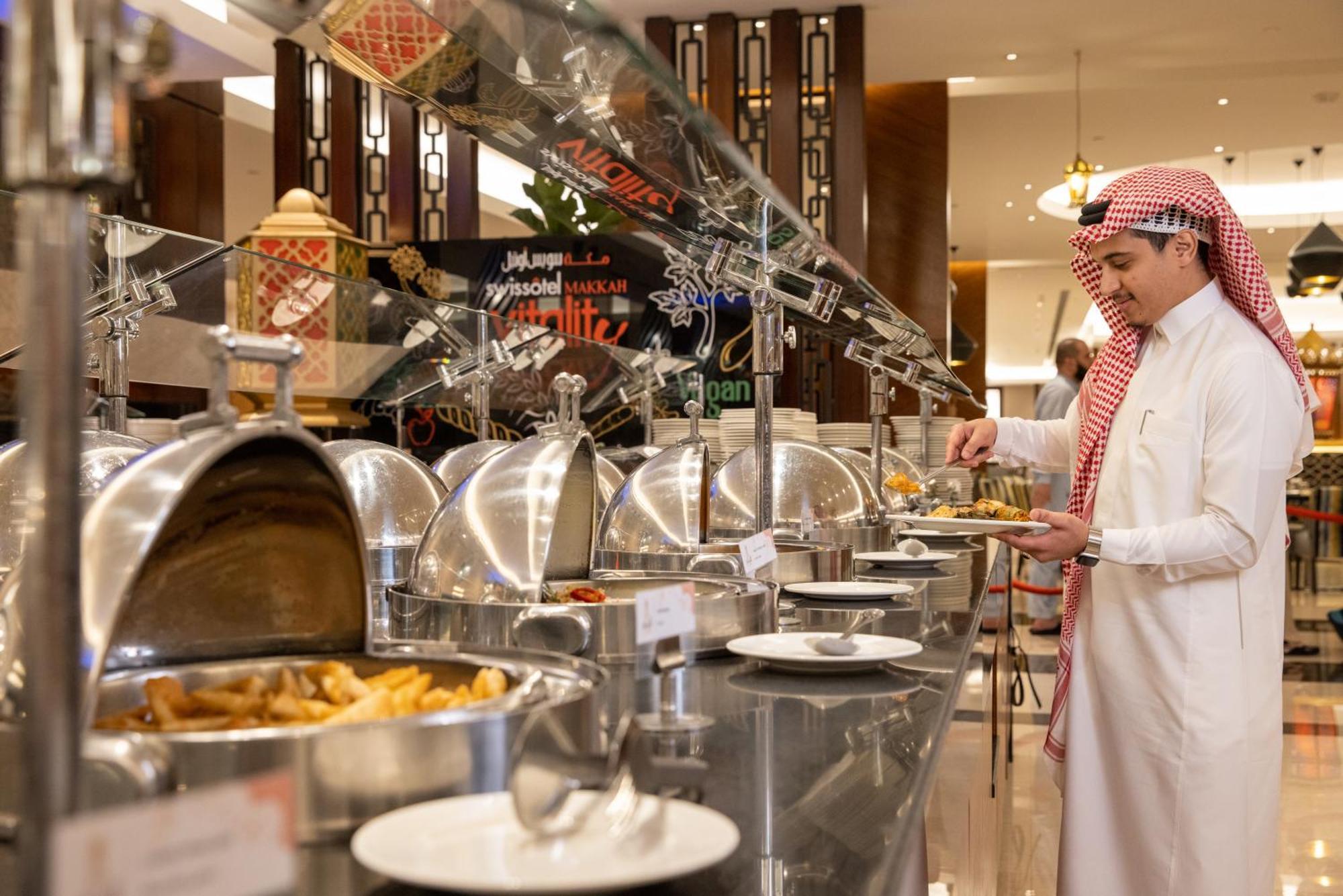 Swissotel Makkah Mecca Exterior photo The photo shows a buffet-style dining setup. A man dressed in traditional attire is standing in front of a row of shiny silver food warmers. He appears to be selecting food from the buffet. The background has decorative elements, and you can see vari