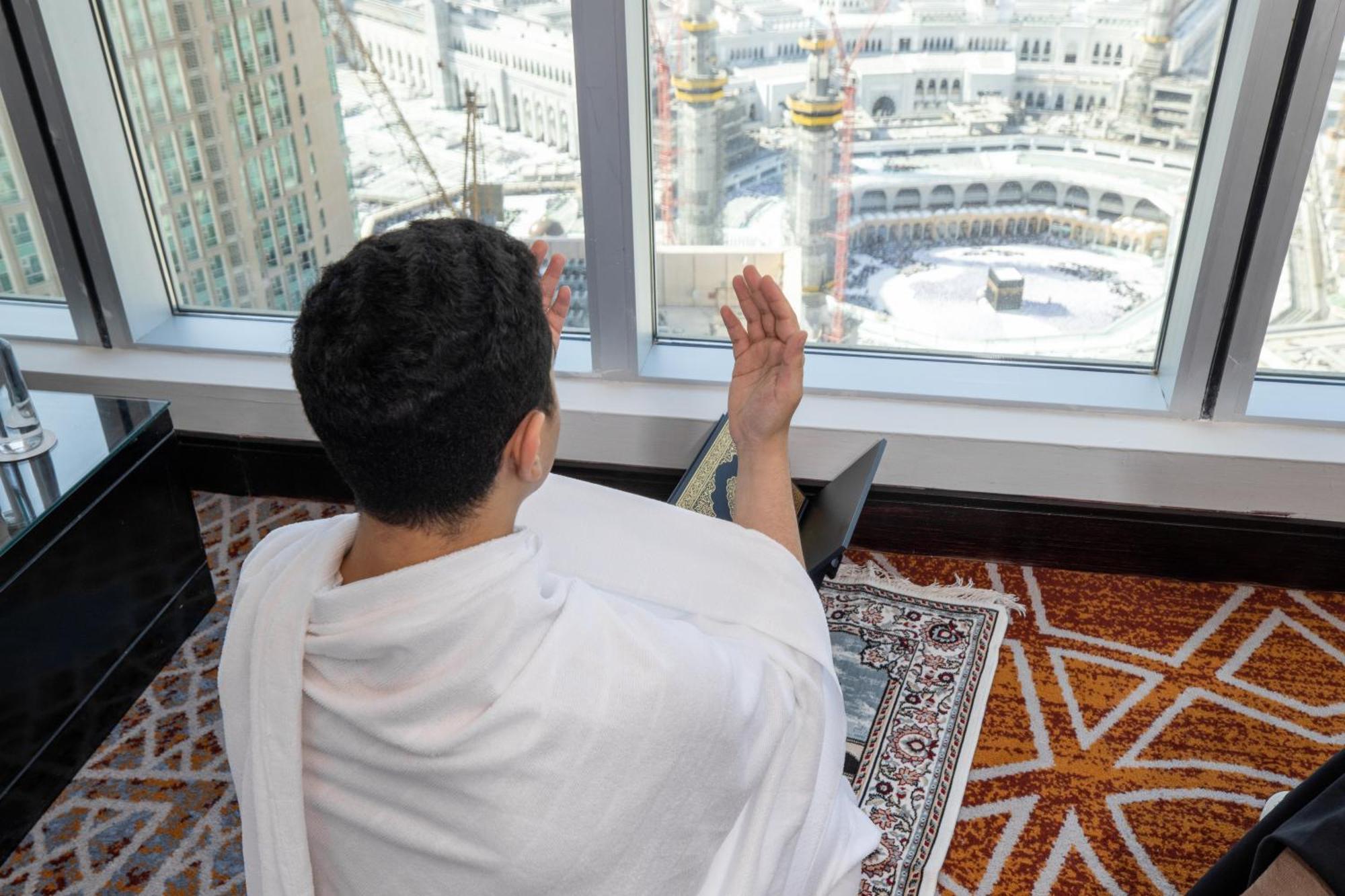 Swissotel Makkah Mecca Exterior photo The photo shows a person in traditional clothing, likely a Ihram, sitting in a room with a large window that overlooks a holy site, presumably the Kaaba in Mecca. The individual is engaged in prayer, raising their hands in a gesture commonly associat