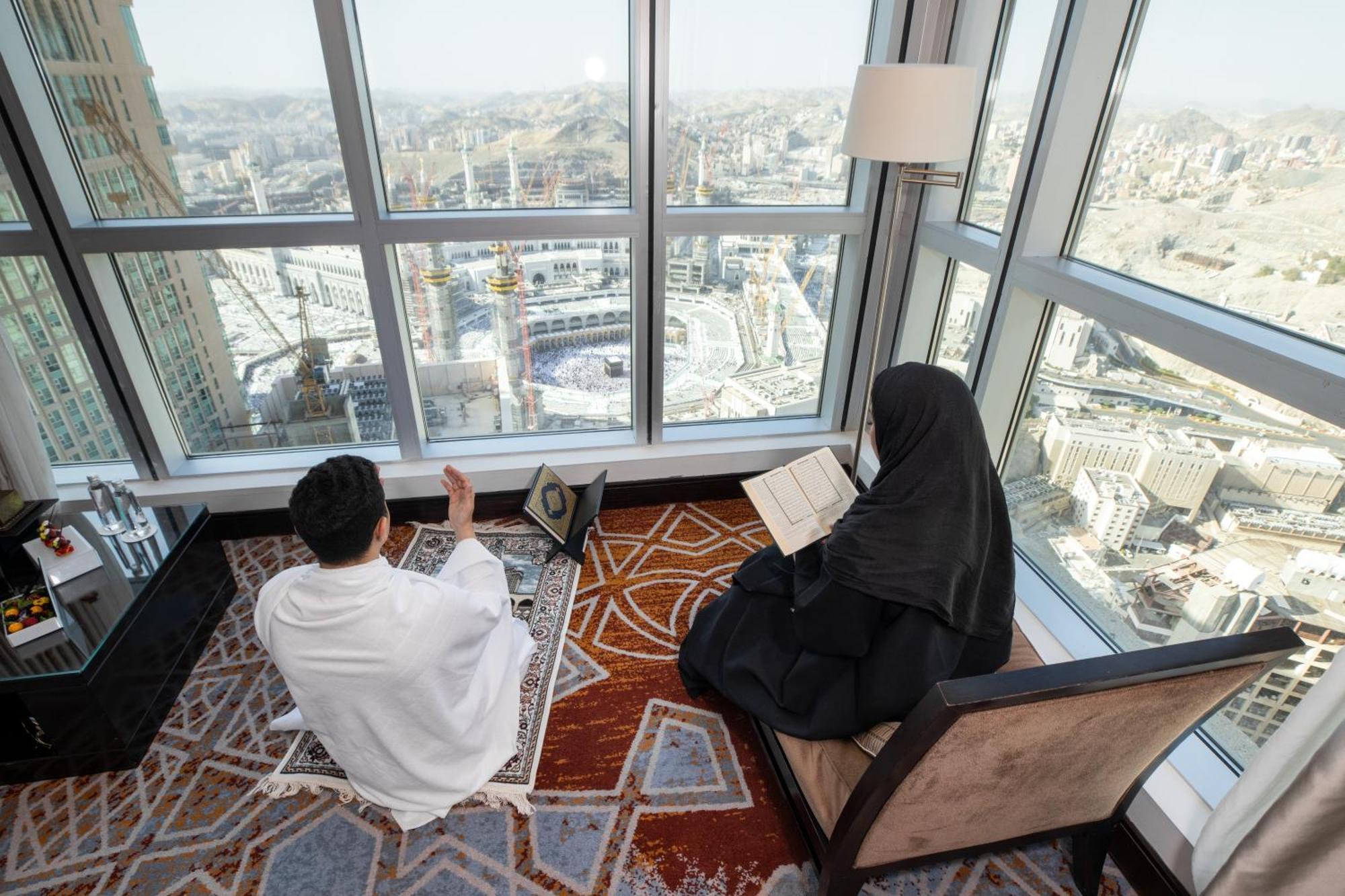 Swissotel Makkah Mecca Exterior photo The photo shows a man and a woman engaged in prayer and reading, respectively, inside a room with large windows offering a view of a cityscape. The man is dressed in traditional attire, facing a direction, possibly in a state of supplication. The wom