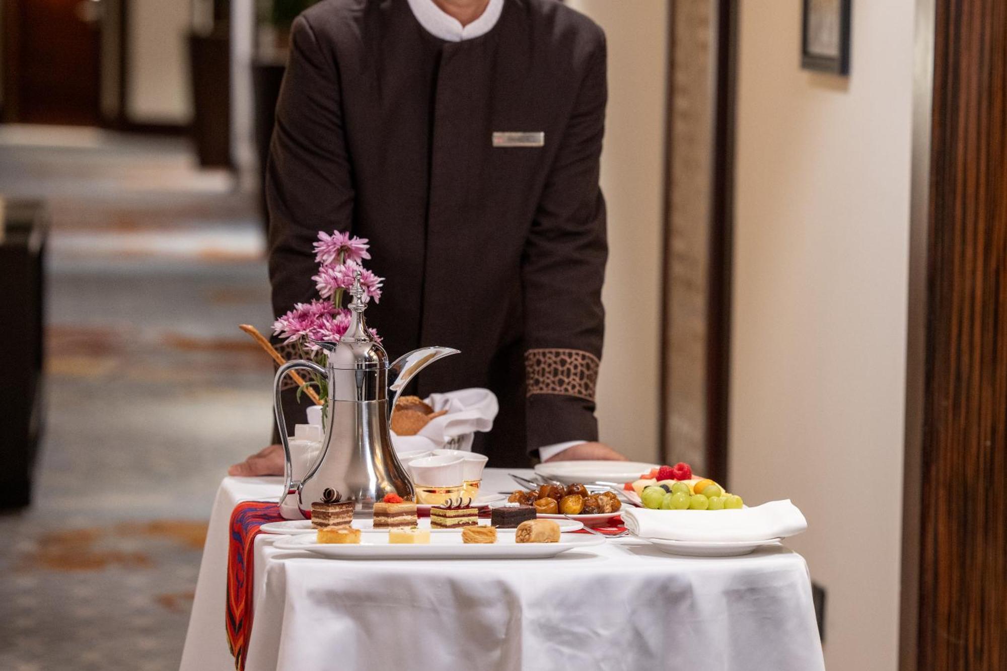 Swissotel Makkah Mecca Exterior photo The photo shows a waiter standing beside a table set with a variety of food and beverages. The table is covered with a white cloth and includes a silver teapot, an assortment of pastries, fruits like grapes and strawberries, and small dishes of sweet