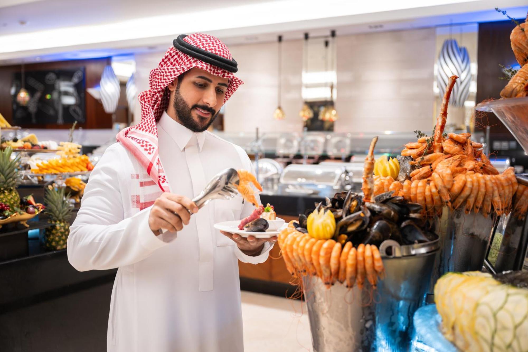 Swissotel Makkah Mecca Exterior photo The photo depicts a man dressed in traditional attire, appearing to be in a restaurant or buffet setting that features a lavish display of seafood. He is using tongs to serve food onto his plate from a large assortment of seafood, which includes item