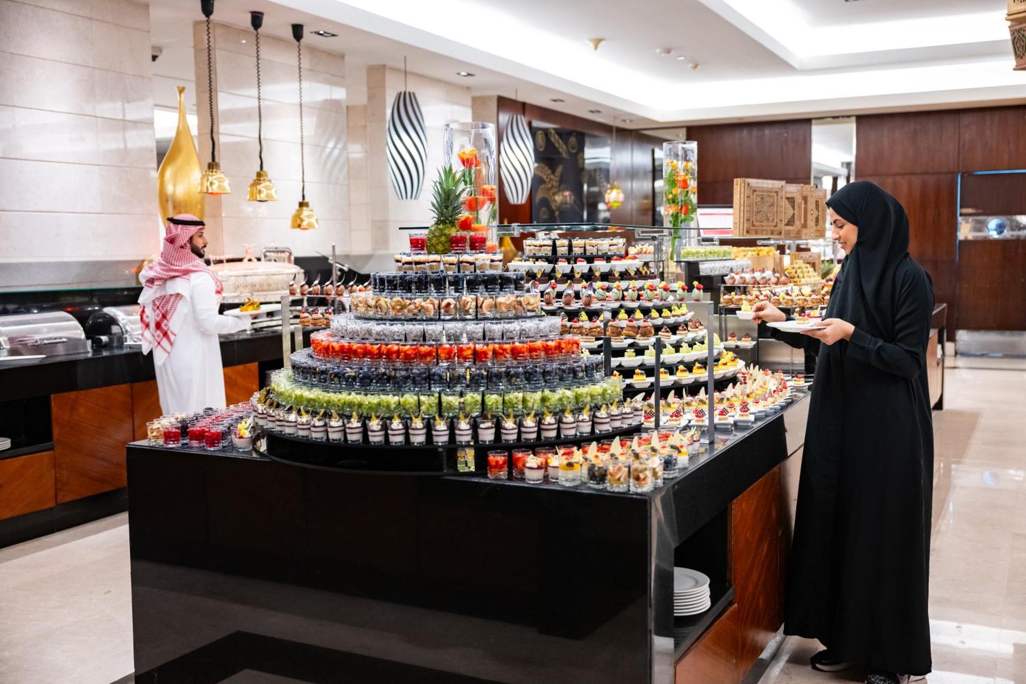 Swissotel Makkah Mecca Exterior photo The photo shows a lavish buffet setup in a dining establishment. There is a large, multi-tiered display of desserts and colorful treats, including various fruits and confectioneries. A woman dressed in a black abaya is seen selecting food from the di