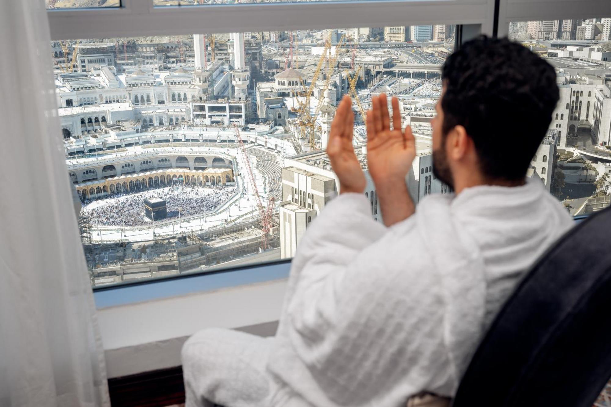 Swissotel Makkah Mecca Exterior photo The photo shows a person wearing a traditional white garment, seated by a large window with a view of a significant religious site. They appear to be in an attitude of prayer or supplication, looking out towards a vast area filled with many people. T