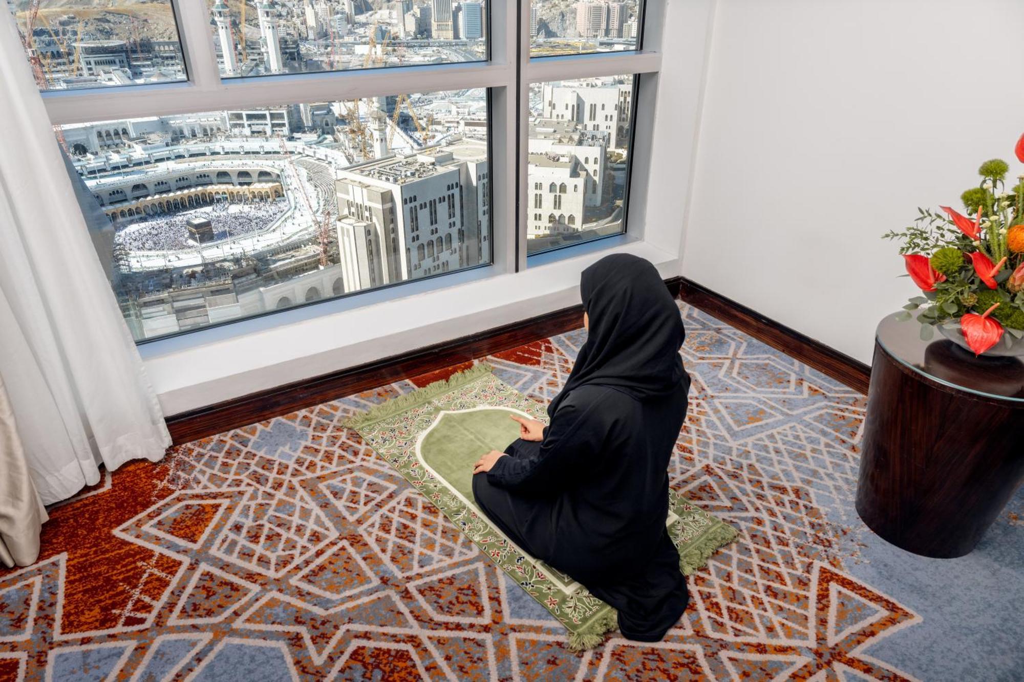 Swissotel Makkah Mecca Exterior photo The photo shows a person sitting on a prayer mat in a room, facing a large window that provides a view of a bustling city. The setting appears to be in a hotel or high-rise building, with the iconic structure of the Kaaba visible in the distance, sug