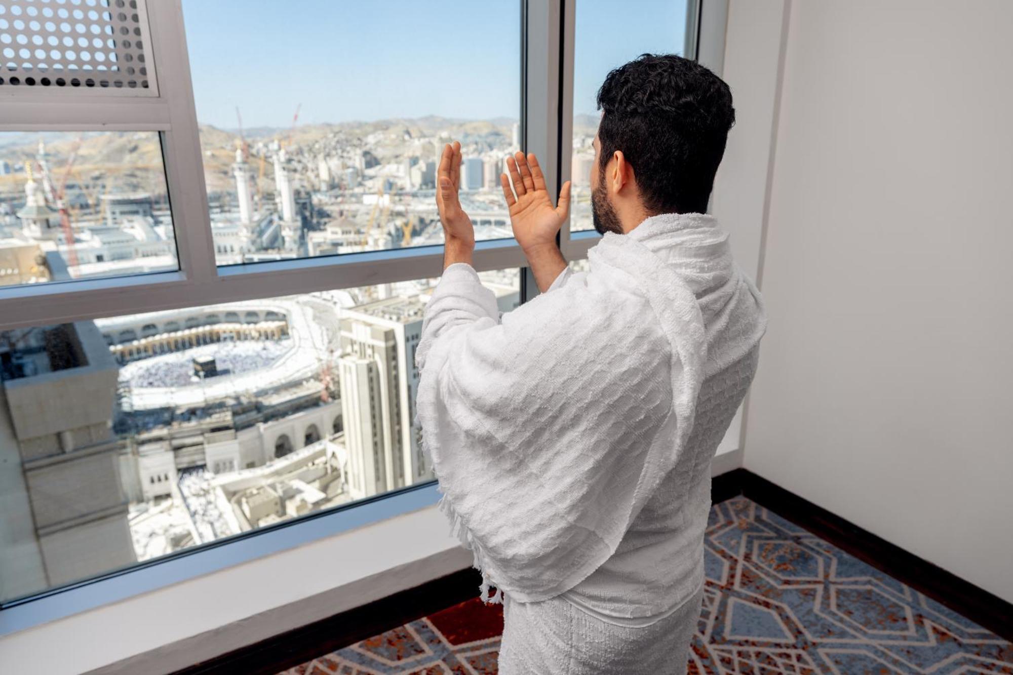 Swissotel Makkah Mecca Exterior photo The photo shows a man dressed in traditional attire, likely a white ihram garment, standing by a window. He appears to be facing towards the city of Mecca, specifically towards the Kaaba, as he is raising his hands in a gesture of prayer or supplicat