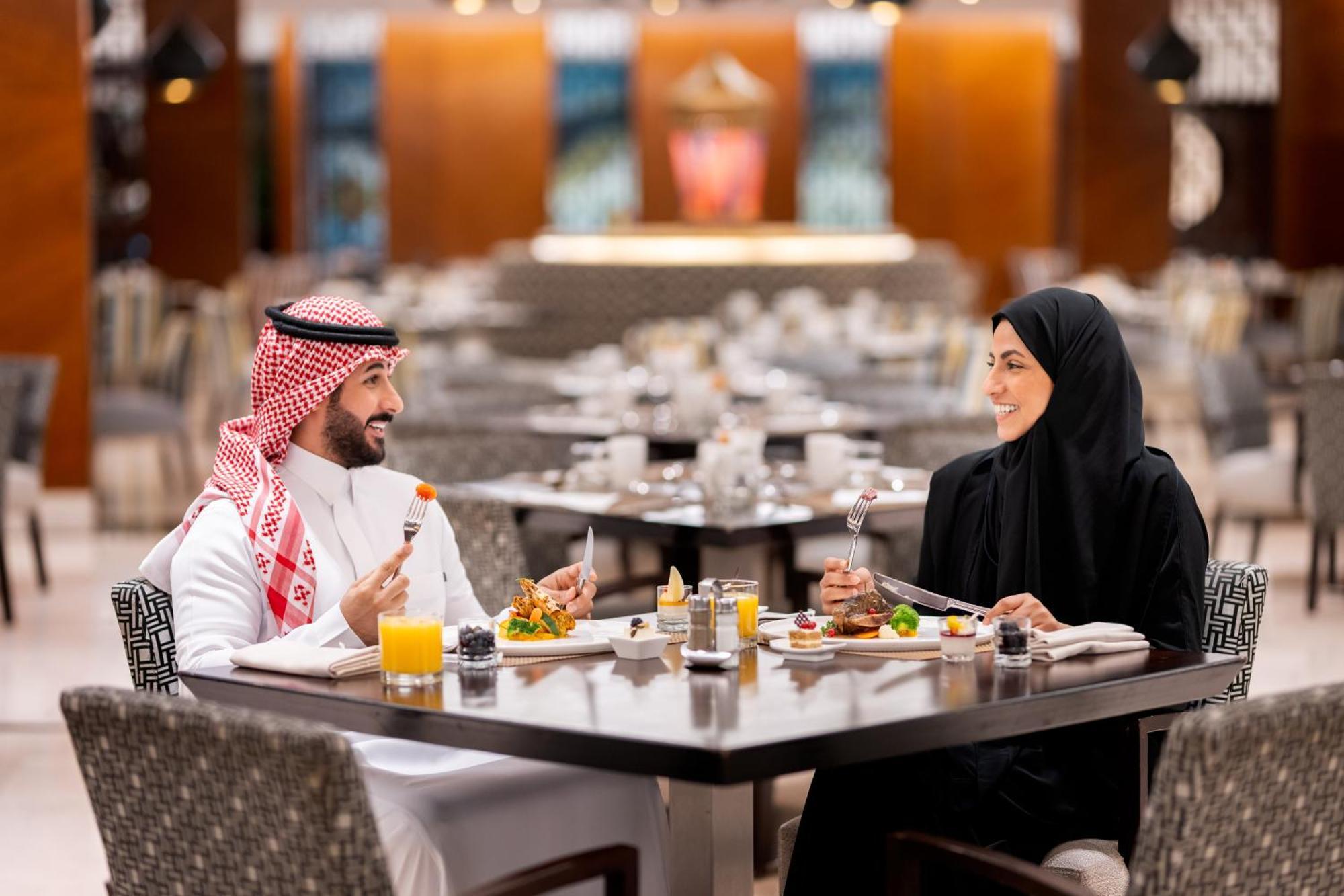 Swissotel Makkah Mecca Exterior photo The photo depicts a couple dining together at a restaurant. The man is dressed in traditional Saudi attire, which includes a white thobe and a red-and-white checked ghutrah (headscarf). The woman is wearing a black abaya and hijab. They are seated at