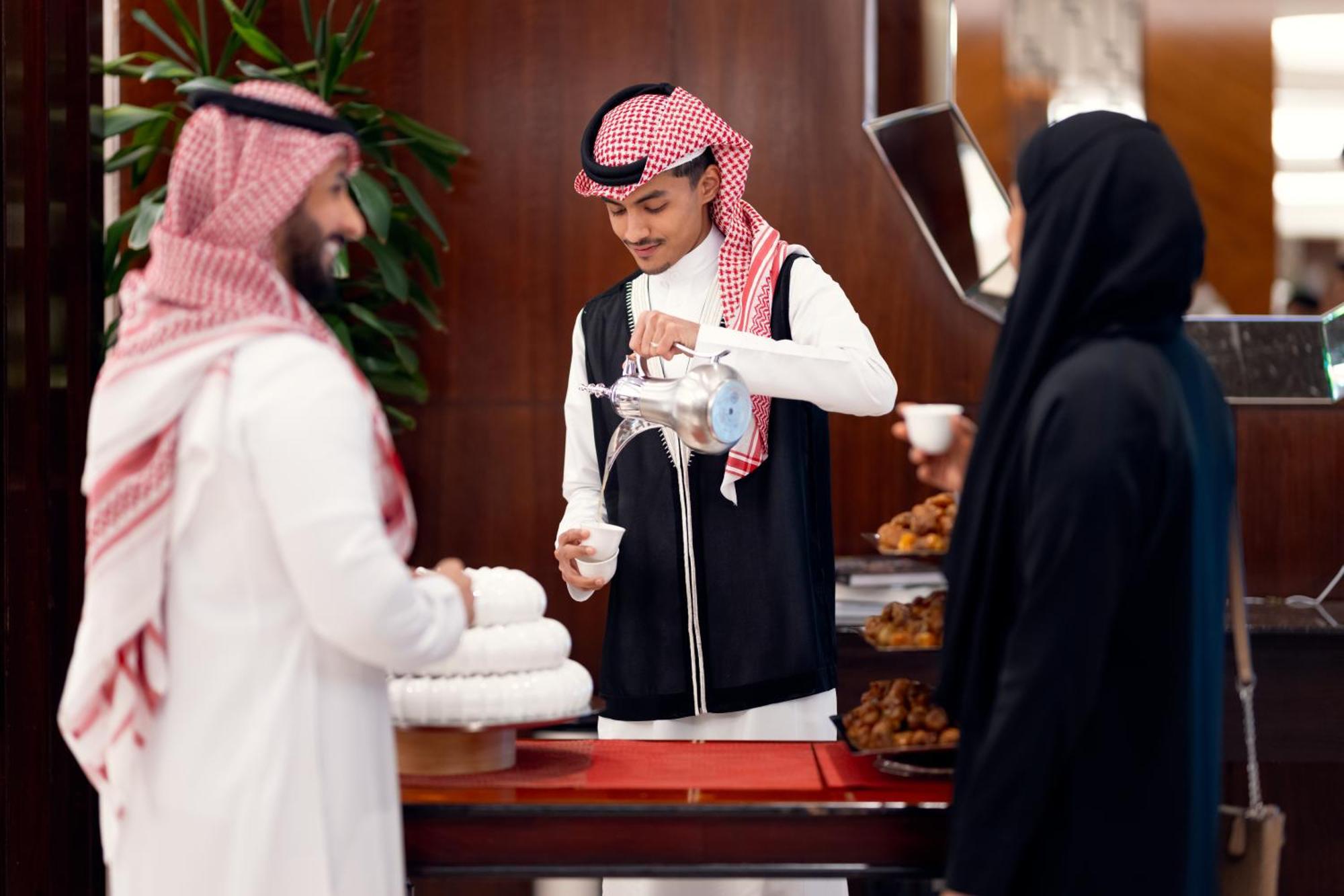 Swissotel Makkah Mecca Exterior photo The photo depicts a scene in a setting that resembles a formal gathering or event. In the foreground, a man in traditional attire, wearing a white thobe with a red and white checked headscarf, is pouring a beverage from a silver coffee pot into a sma