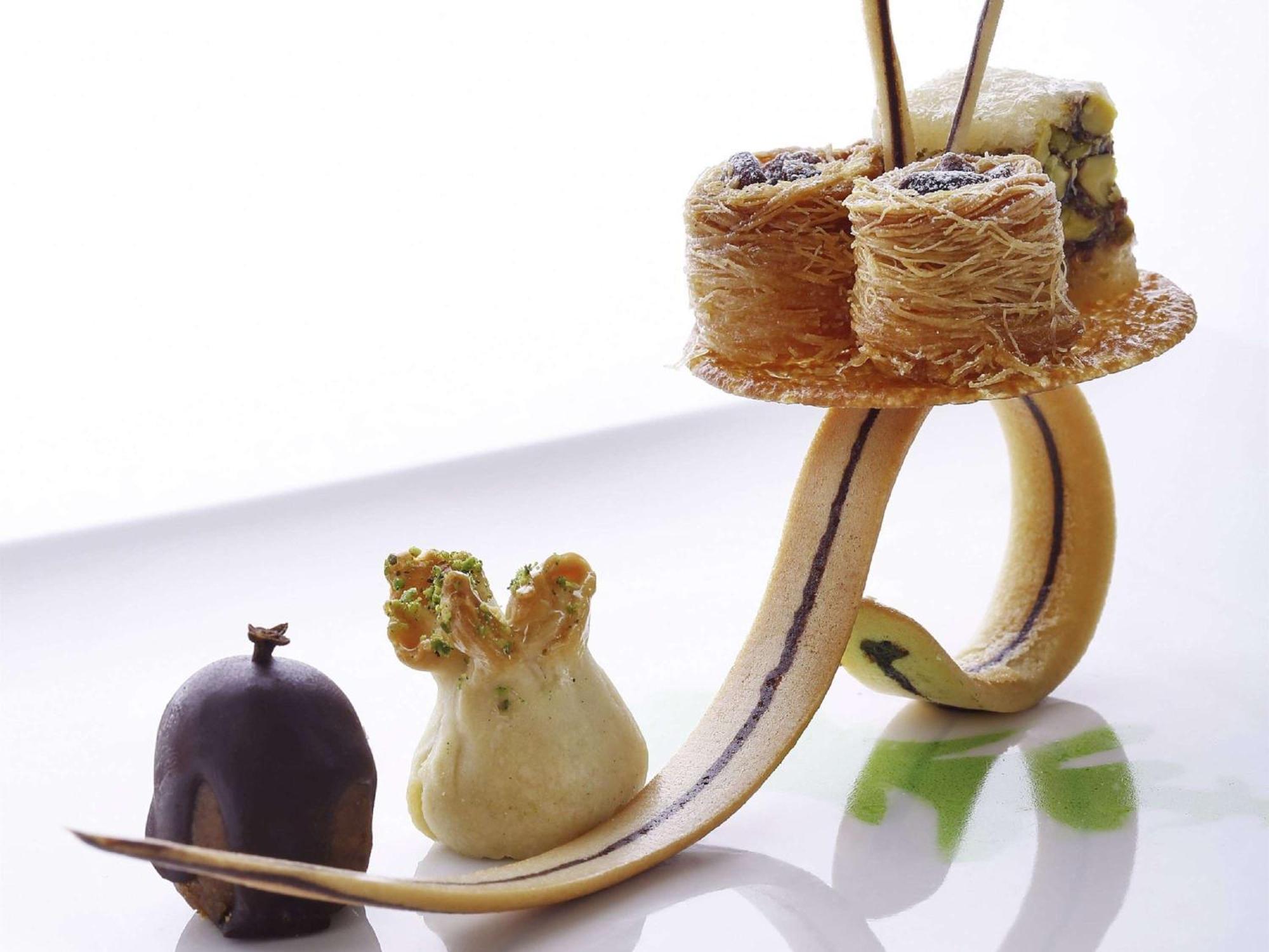 Swissotel Makkah Mecca Exterior photo The photo shows an artistic dessert presentation. It features a curved, elongated base that resembles a banana peel or similar shape. On this base, there are several small desserts arranged creatively. 

1. **Baklava**: There are pieces of baklava, w