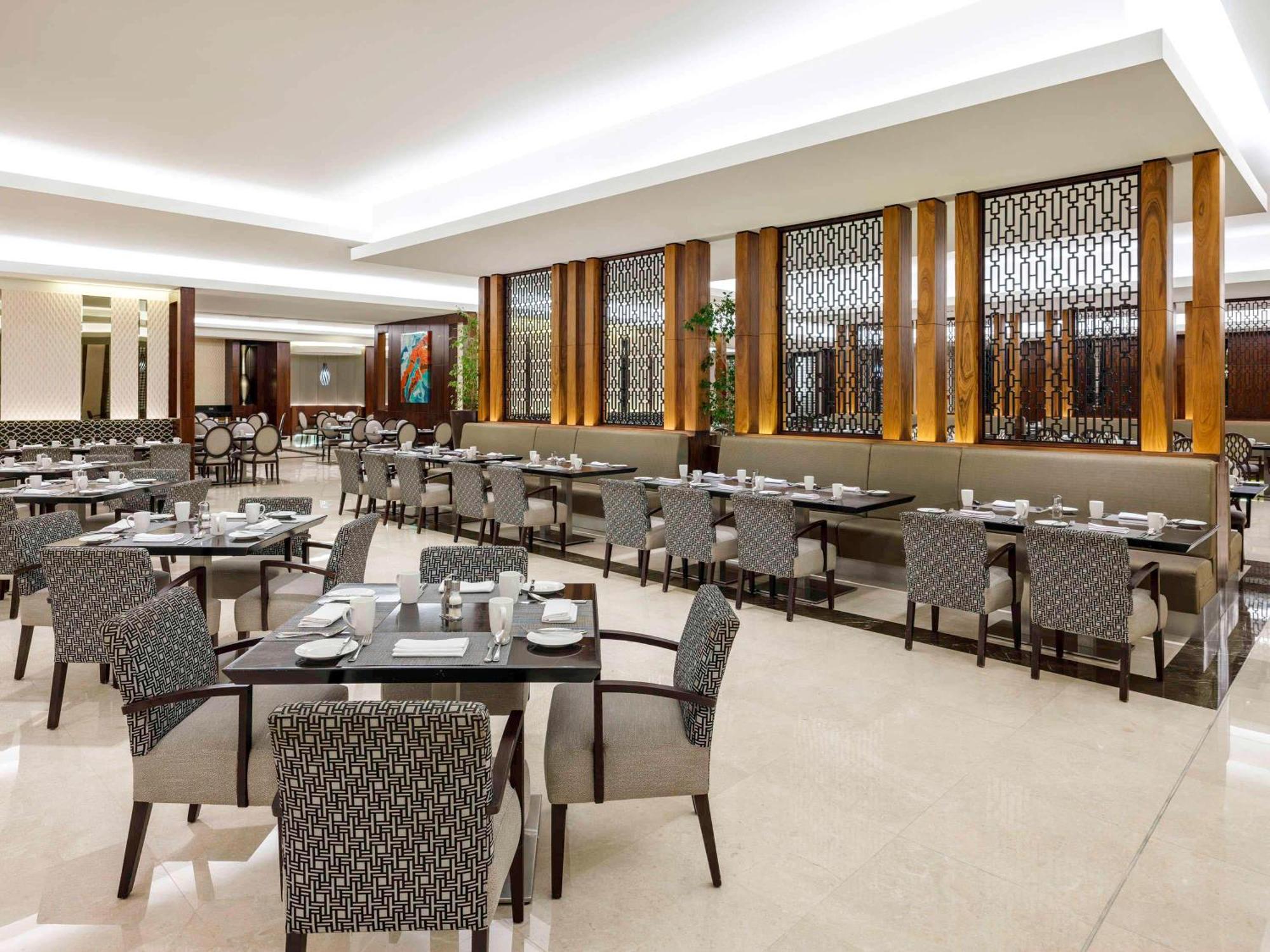 Swissotel Makkah Mecca Exterior photo The photo shows a modern dining area in a restaurant. The space features multiple tables covered with white tableware and neatly arranged chairs. The chairs have a patterned fabric, and the walls are decorated with wooden accents and a decorative scr