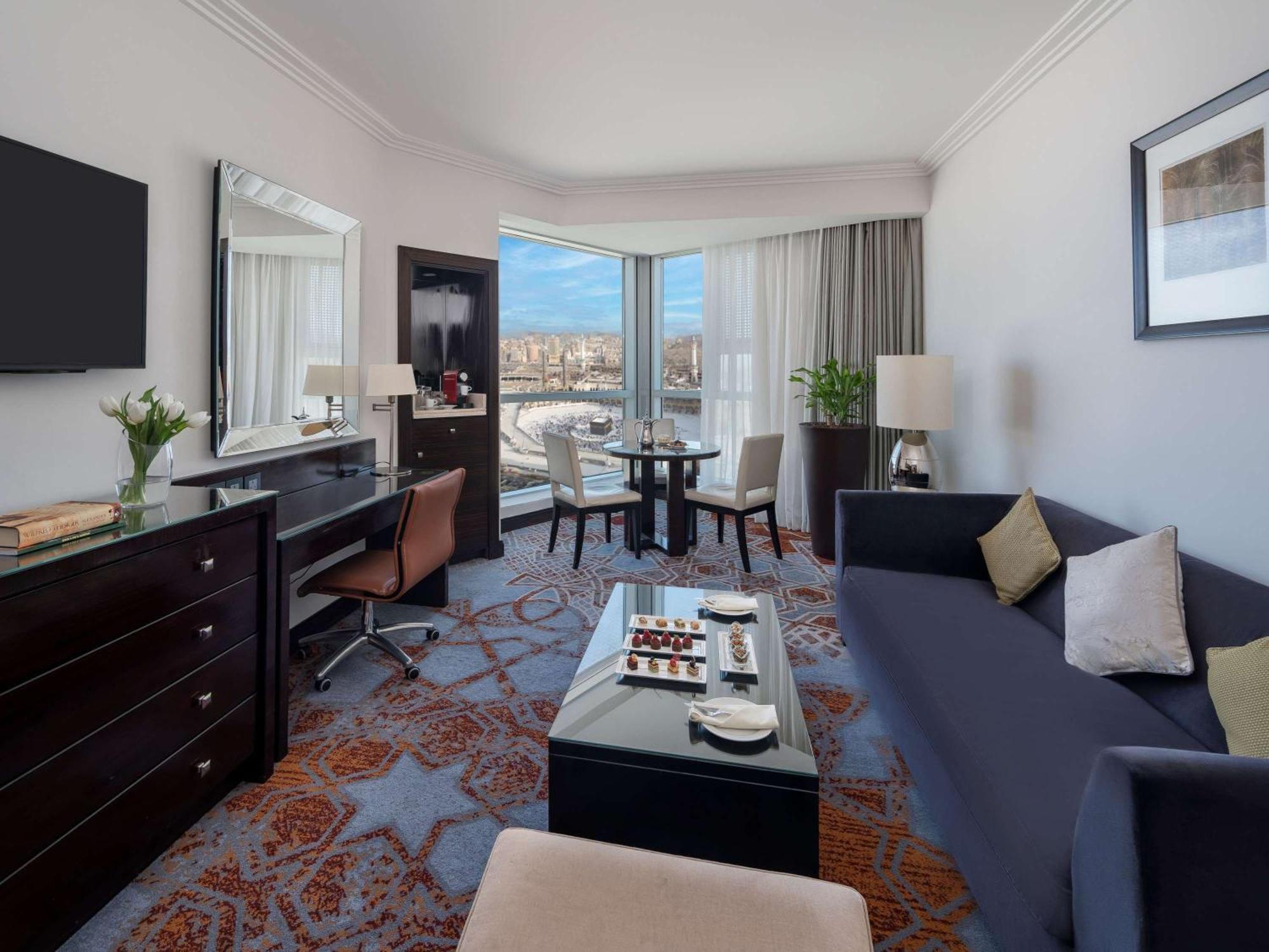 Swissotel Makkah Mecca Exterior photo The photo shows a hotel room featuring a modern and stylish design. There is a comfortable dark blue sofa and a coffee table in the center, along with light-colored accent pillows. Nearby, a small dining table with chairs is positioned near large win