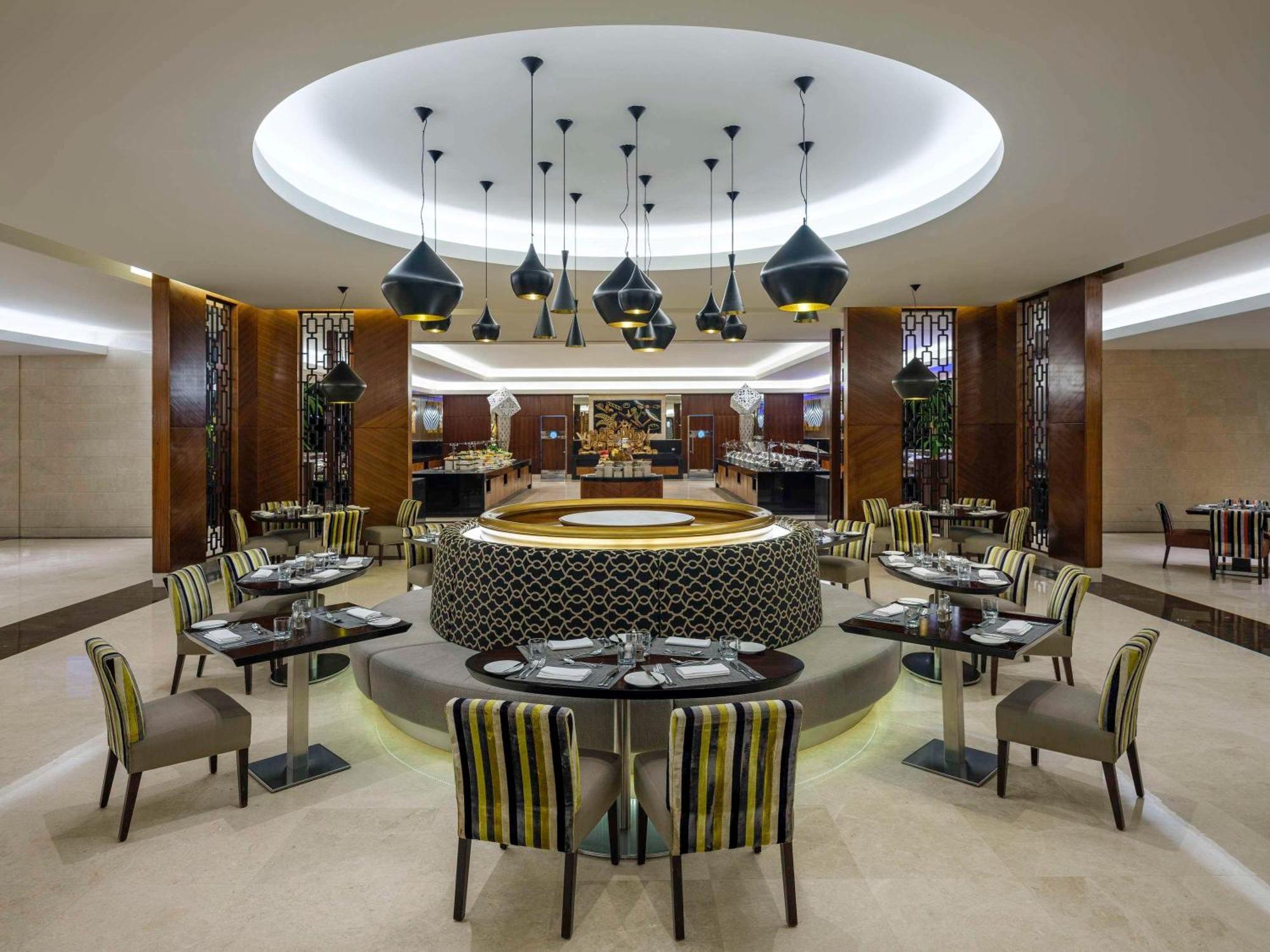 Swissotel Makkah Mecca Exterior photo The photo depicts a modern restaurant interior. The design features a circular dining area with several tables and chairs arranged around a central circular space. The tables are set with cutlery and plates, and some have striped green and brown chai