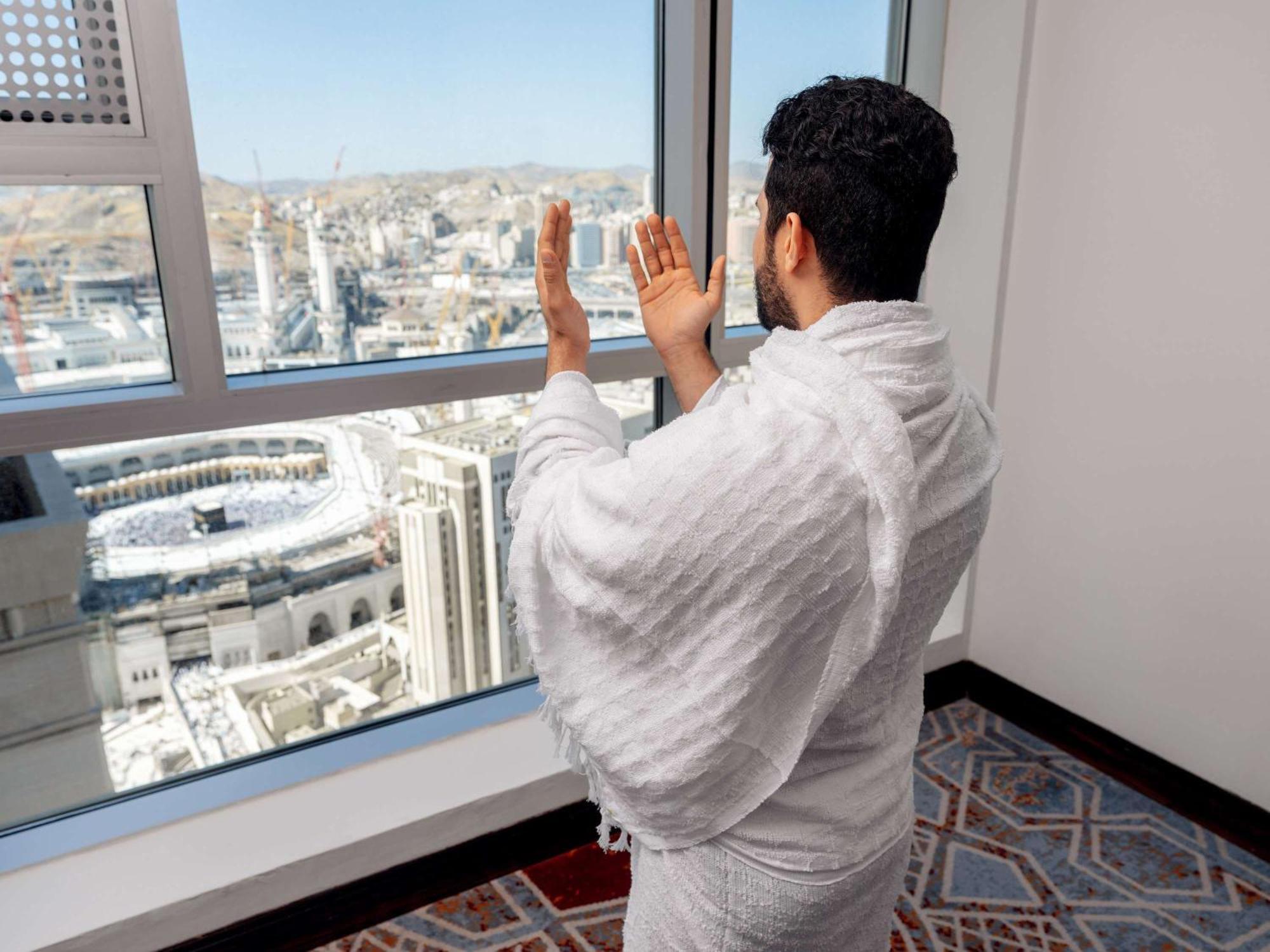 Swissotel Makkah Mecca Exterior photo The photo shows a person standing by a window in a room, wearing traditional white clothing. The individual appears to be in a prayer or supplication pose, looking out at a cityscape that includes a view of the Kaaba, a significant religious site loc