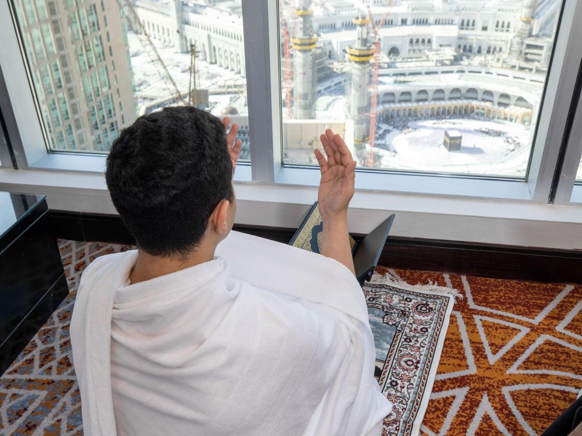 Swissotel Makkah Mecca Exterior photo The photo shows a person wearing a white garment, likely a Ihram, which is typically worn during the Hajj pilgrimage. They are seated in a room, facing a large window that offers a view of the Kaaba in Mecca. The individual appears to be engaged in p