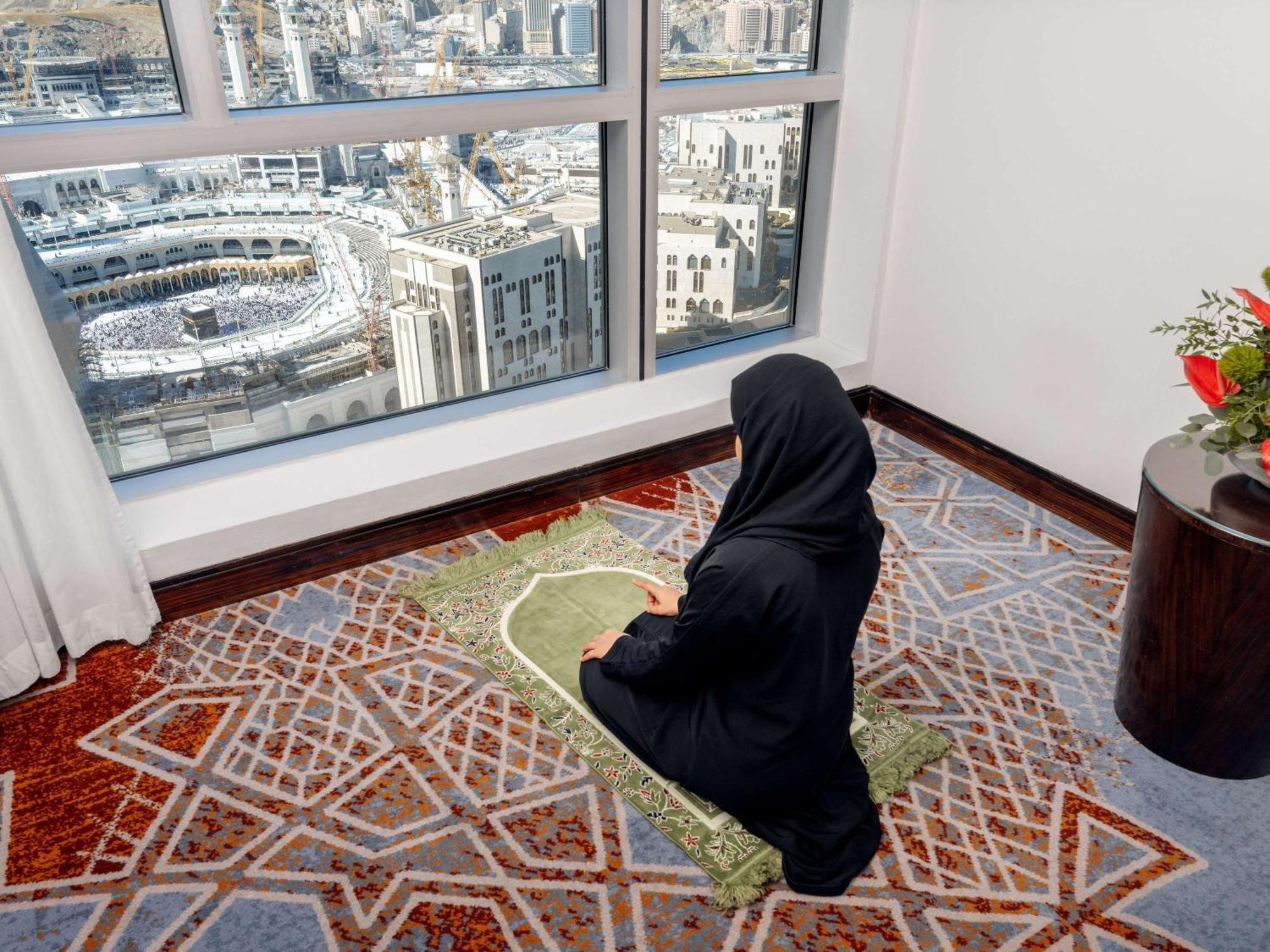 Swissotel Makkah Mecca Exterior photo The photo shows a person sitting on a prayer mat in front of large windows that offer a view of a city, likely Mecca, as indicated by the architectural features visible. The individual is wearing a black garment, which is typical attire in some cultu