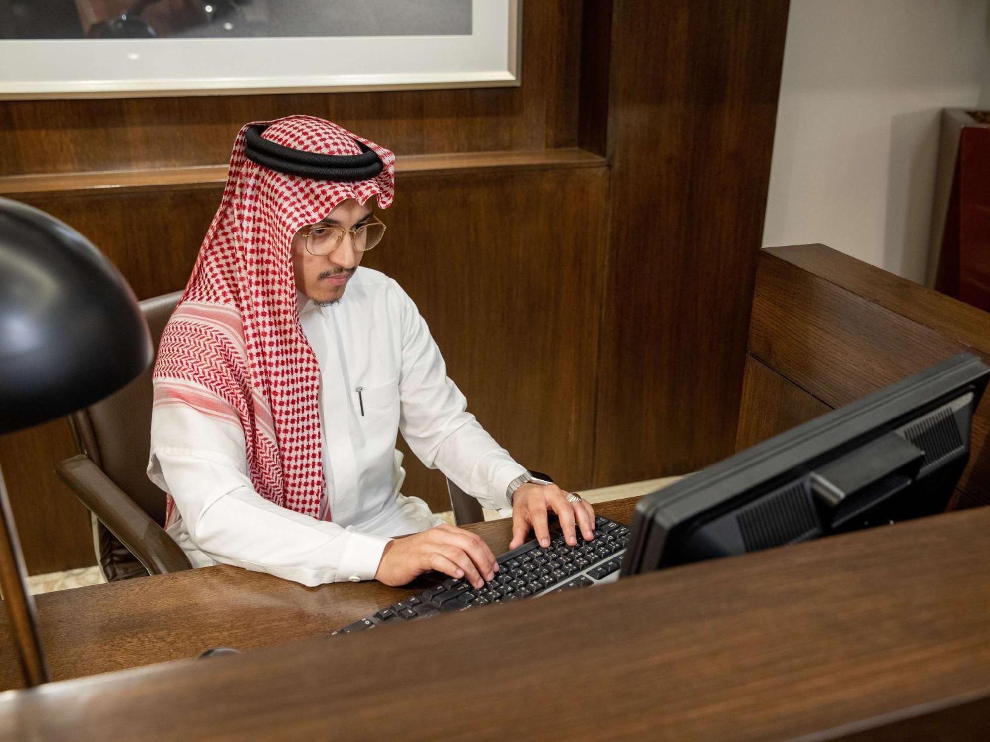 Swissotel Makkah Mecca Exterior photo The photo shows a man sitting at a desk, working on a computer. He is wearing traditional attire, including a white thobe and a red-and-white checked shemagh headdress. The setting appears to be an office, characterized by wooden furnishings and an o