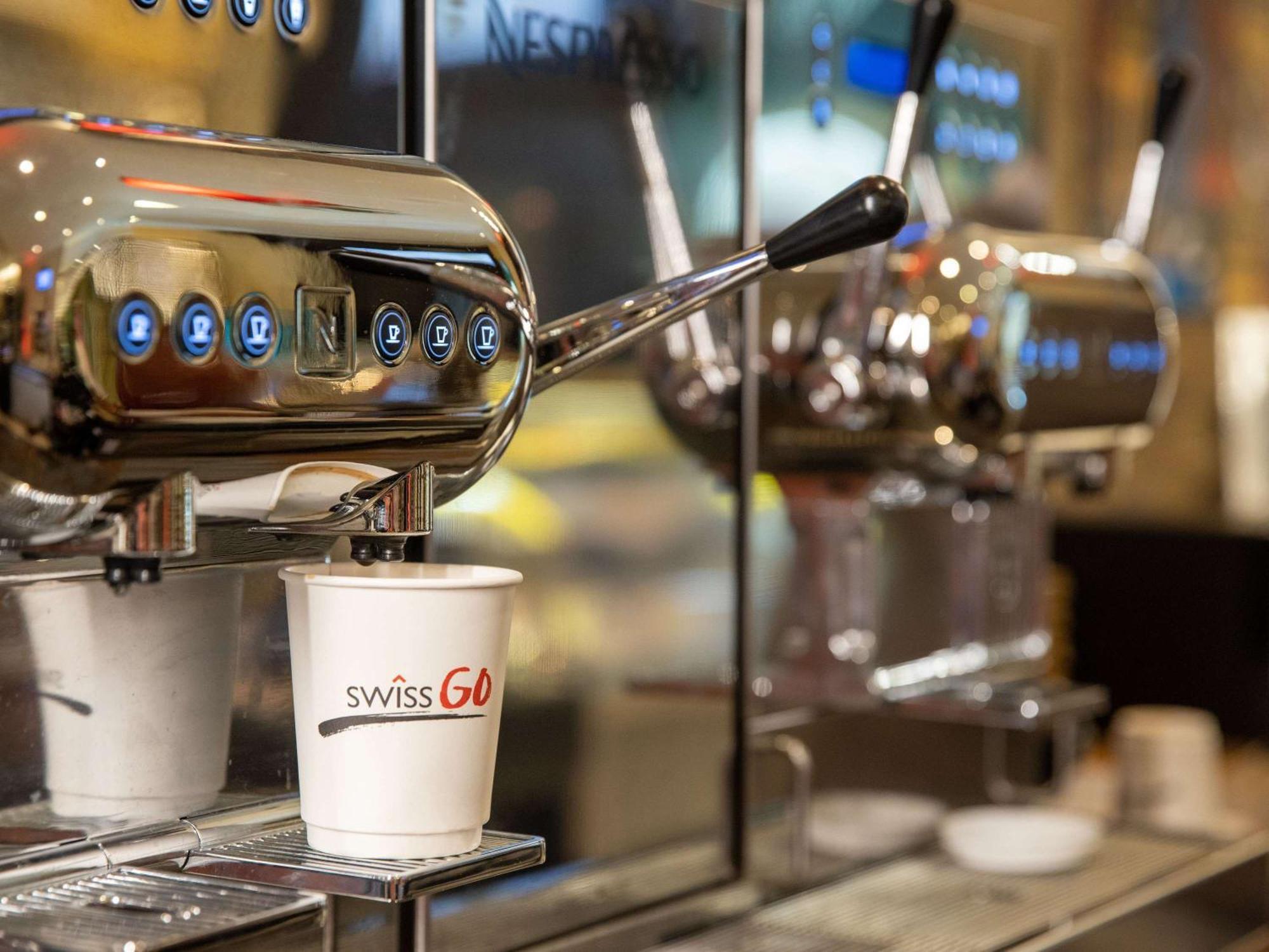 Swissotel Makkah Mecca Exterior photo The photo shows a coffee machine setup designed for making espresso. In the foreground, there is a white paper cup with the logo "swissGo" displayed prominently. Behind the cup, you can see a shiny, metallic espresso machine with multiple controls an