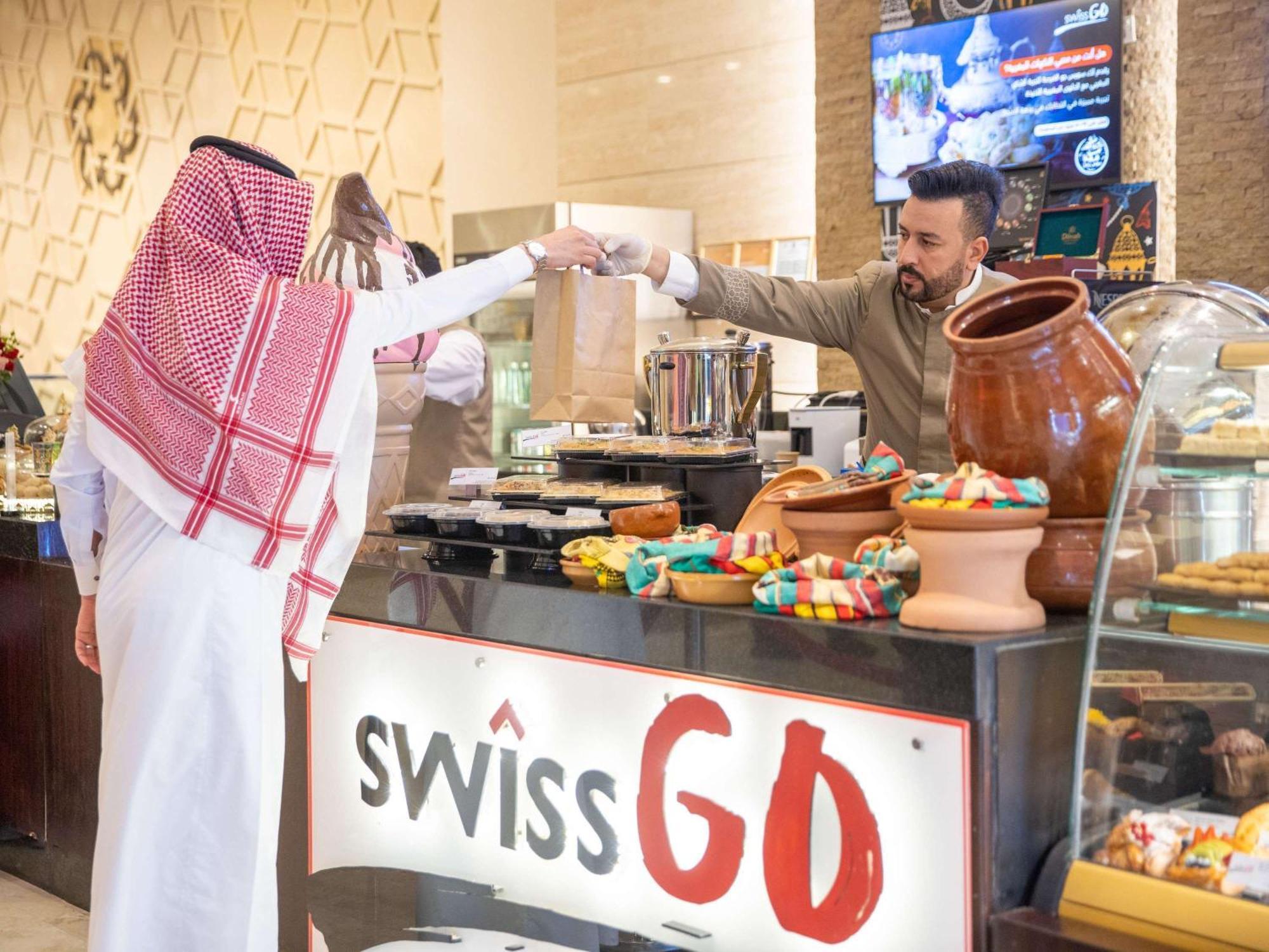 Swissotel Makkah Mecca Exterior photo The photo shows a scene in a restaurant or café called "Swiss GO." In the foreground, a customer wearing traditional attire is interacting with a server behind the counter. The server is handing over a bag or food item. The counter features a variety