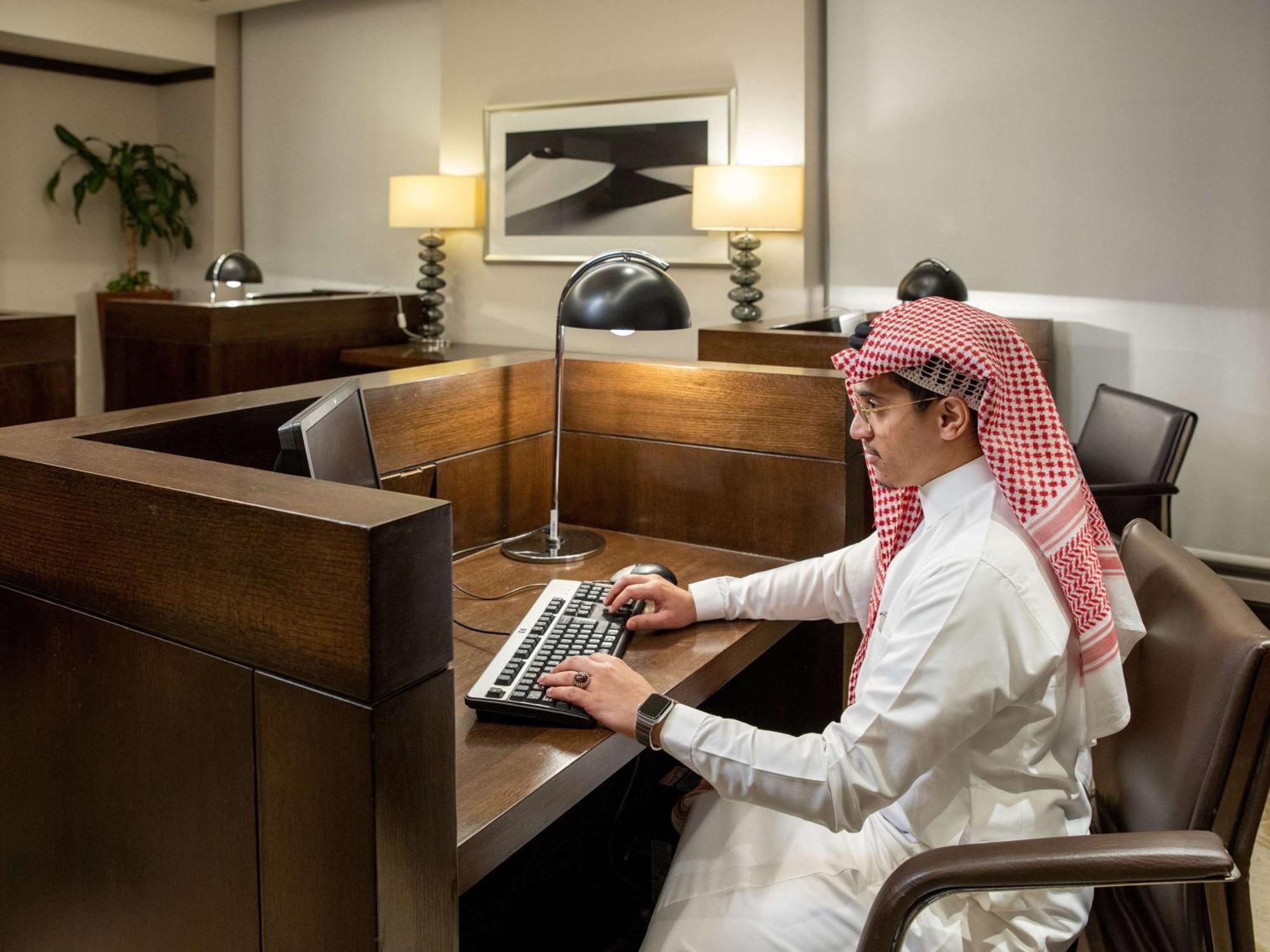 Swissotel Makkah Mecca Exterior photo The photo shows a man seated at a wooden desk in a modern office setting. He is dressed in traditional attire, which includes a white thobe and a red and white checked headscarf. The man is focused on typing on a keyboard, and there is a desk lamp pr