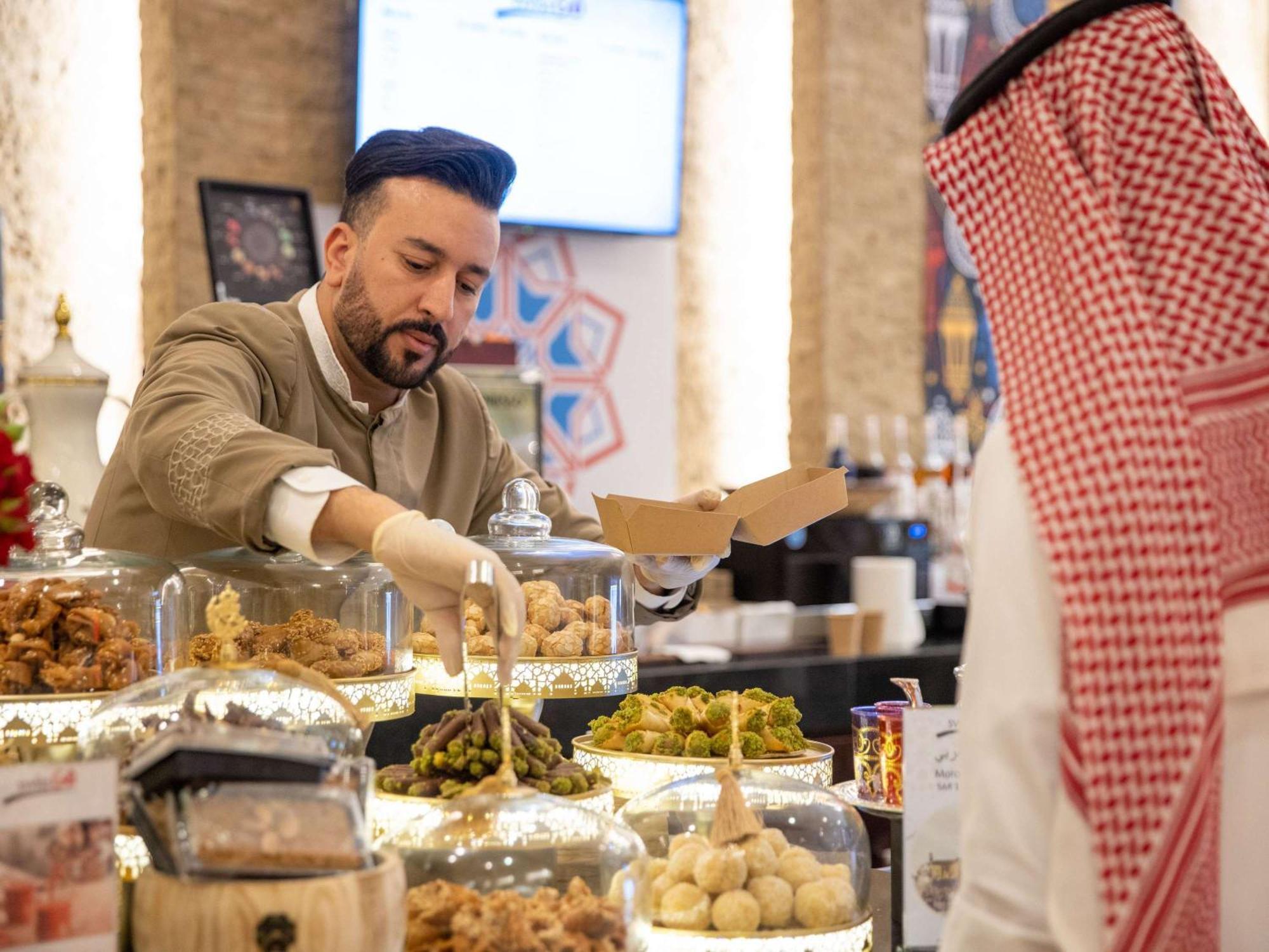 Swissotel Makkah Mecca Exterior photo The photo shows a scene in a food establishment, likely a market or café. A man dressed in a traditional outfit is serving food from a display of various dishes. He is wearing gloves and is placing items into a box for a customer. The display feature