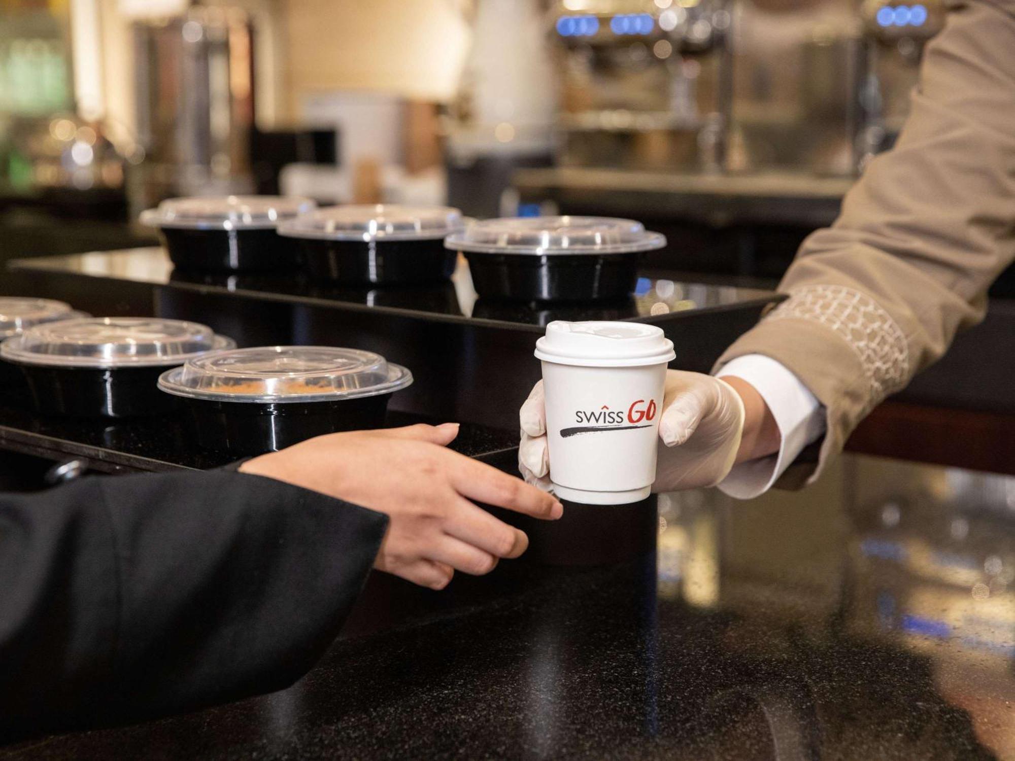 Swissotel Makkah Mecca Exterior photo The image shows a scene at a counter where a person is handing over a cup of coffee with a logo that reads "SwissGo." The person receiving the coffee is wearing a black jacket, while the one serving it is dressed in a beige uniform. In the background