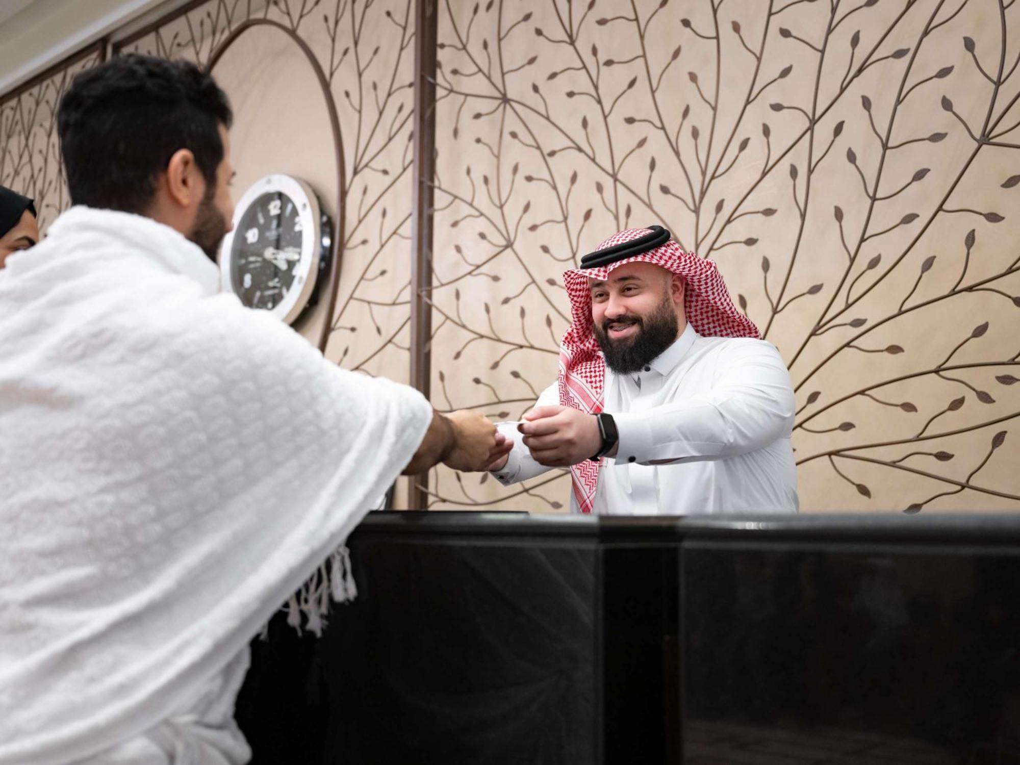 Swissotel Makkah Mecca Exterior photo The image shows two individuals engaged in a handshake. One person, dressed in white traditional attire, appears to be greeting the other, who is wearing a thobe and a traditional headscarf (ghutrah). The background features decorative wall art and a