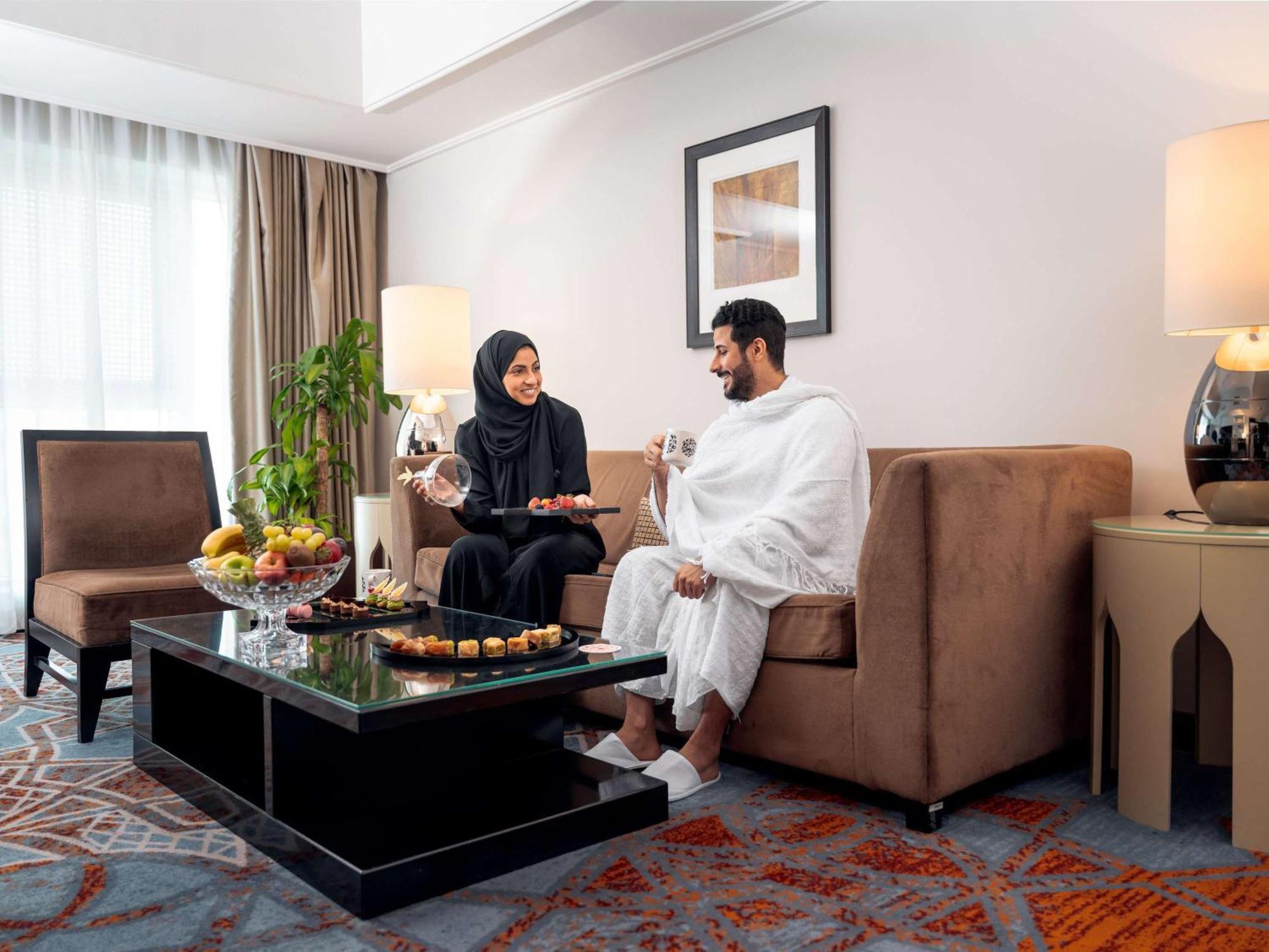 Swissotel Makkah Mecca Exterior photo The photo depicts a comfortable living space where a man and woman are seated together. The man is wearing traditional attire, while the woman is dressed in a black abaya. They appear to be engaged in a pleasant conversation. A coffee table in front 